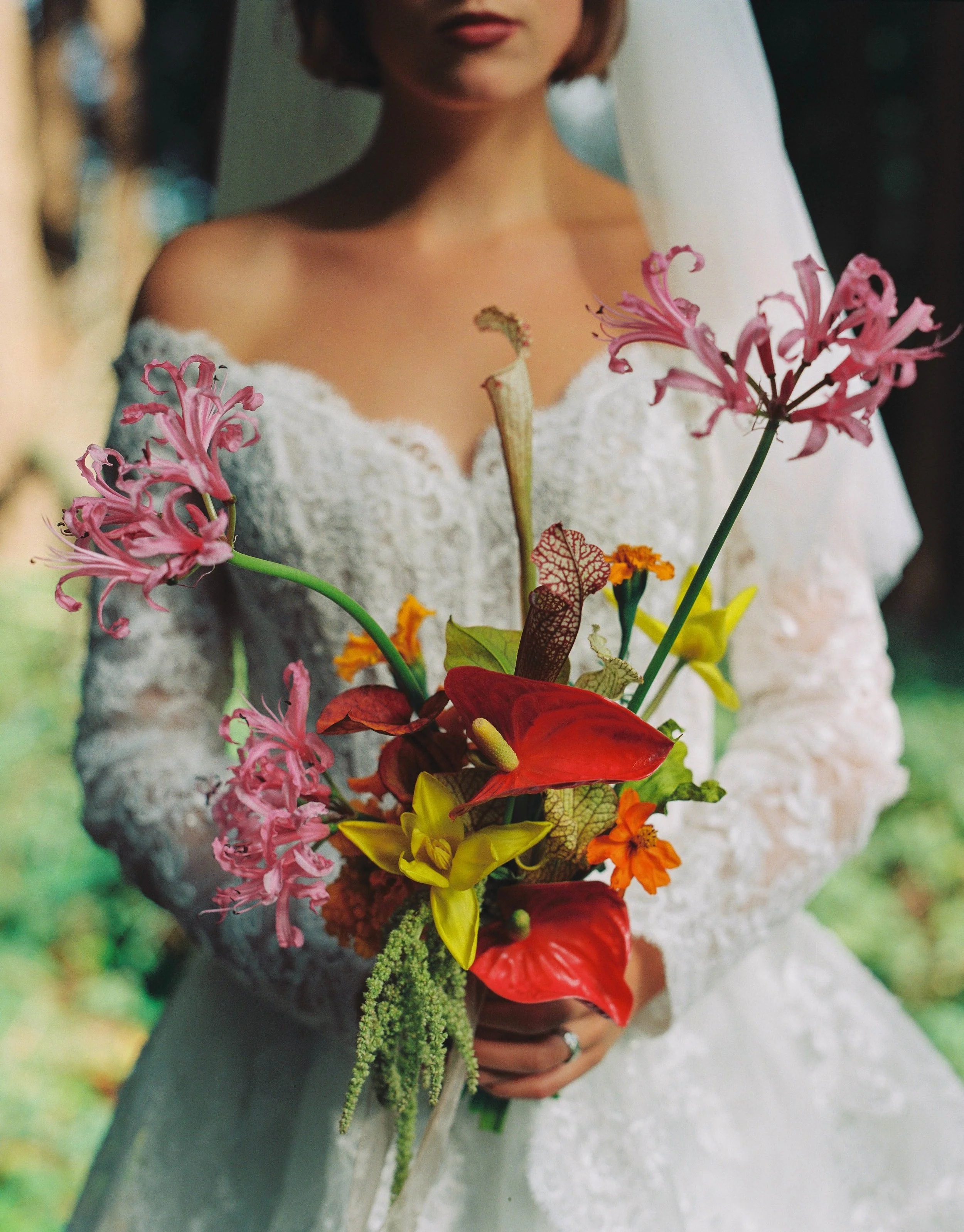 A woman in a white lace wedding dress holding a colorful bouquet of flowers, including pink, red, yellow, and orange blooms, outdoors with greenery in the background.