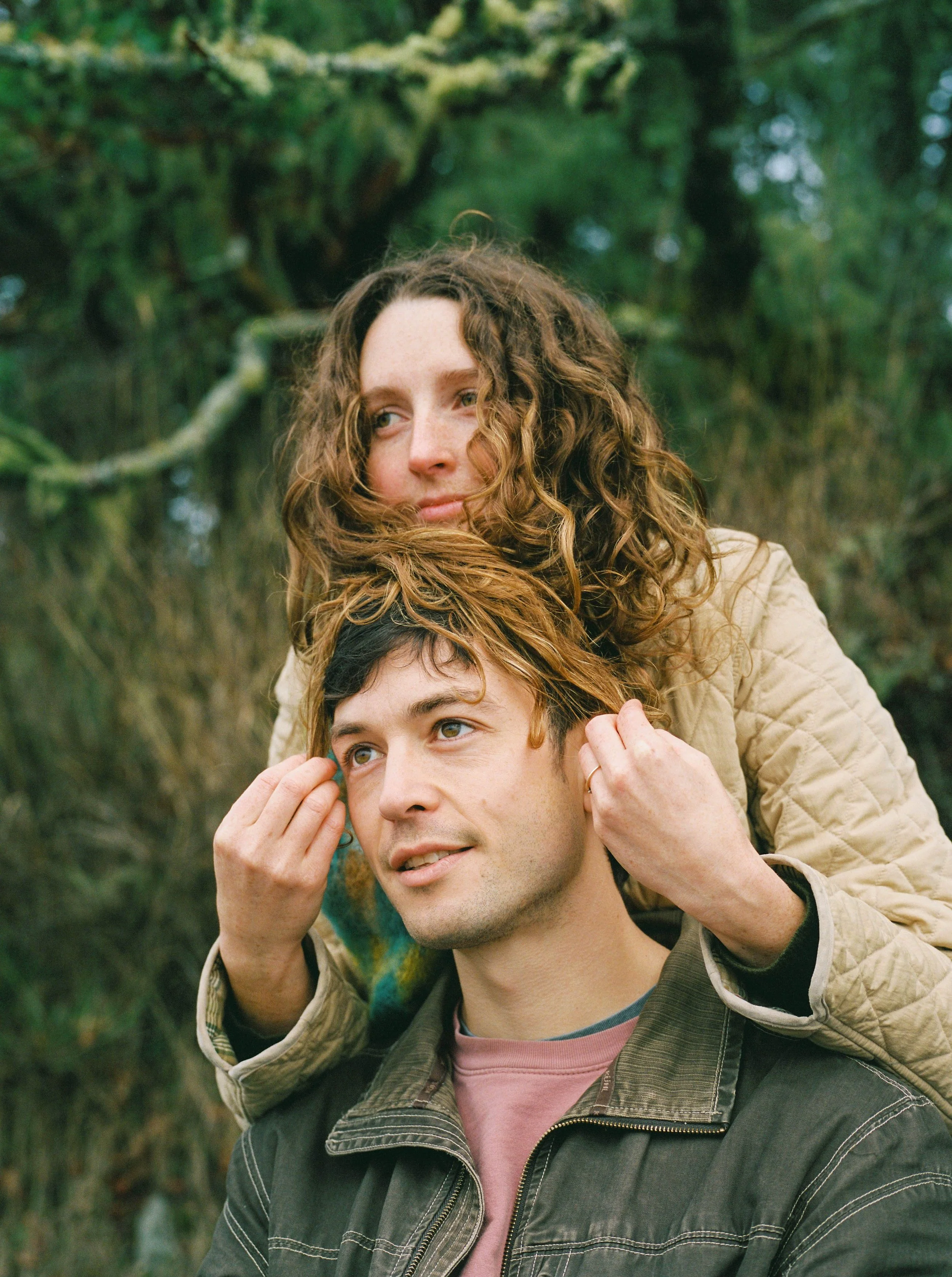 A woman with curly hair resting her head on a man's with short hair while outdoors in a green, wooded area.