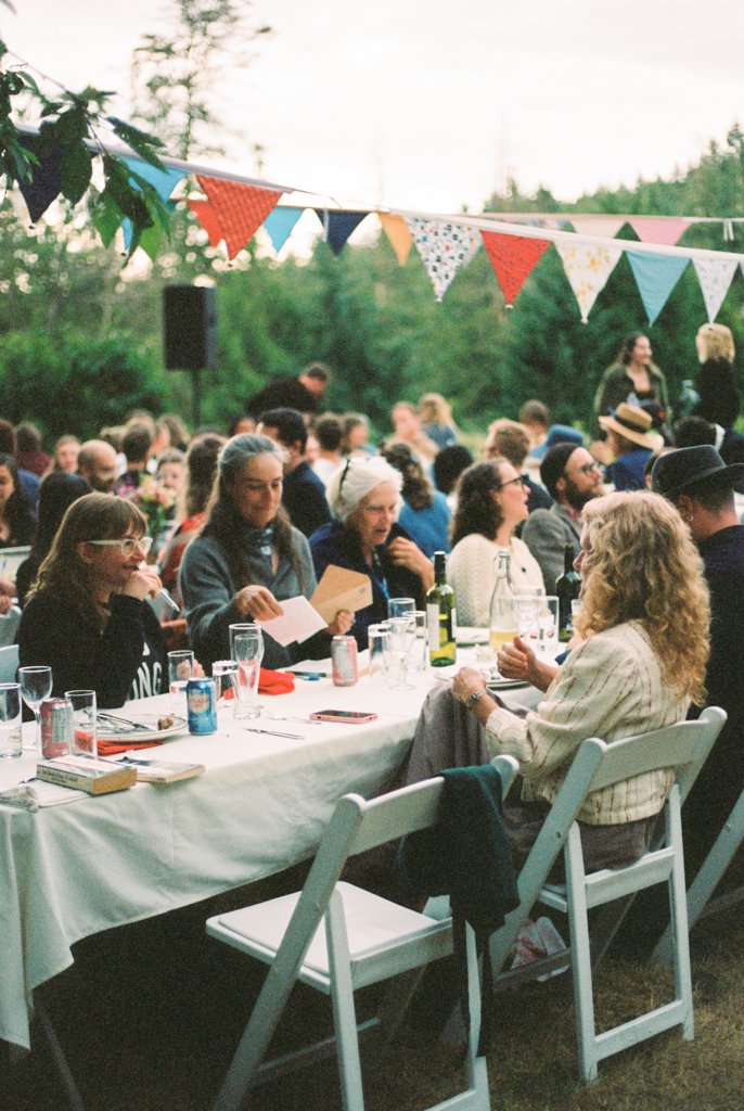 A group of people gathering outdoors under colorful bunting, seated at a long table with drinks, engaged in conversation, with trees and a crowd in the background.