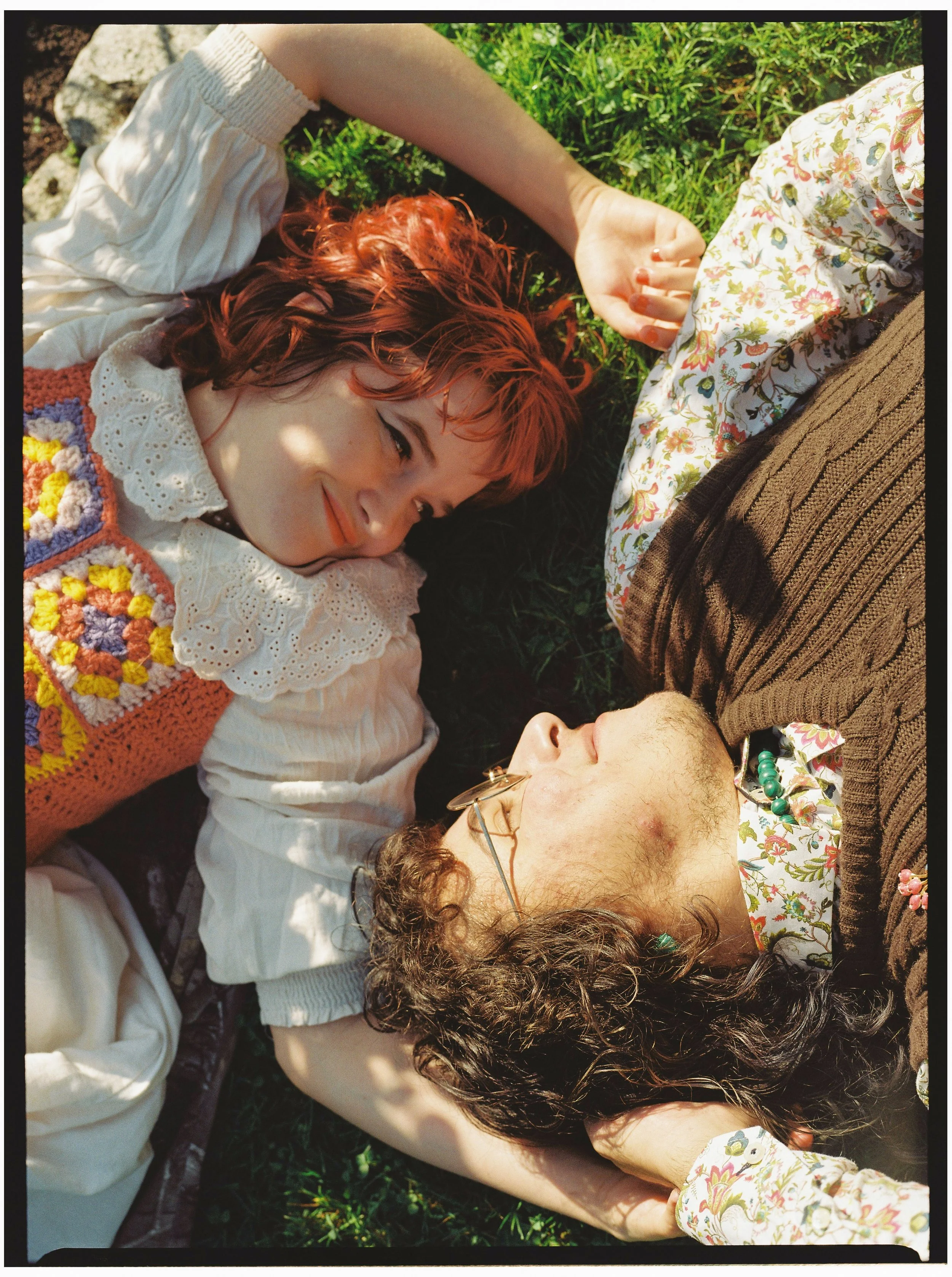 Two women lying on grass, one with curly red hair wearing a white blouse with lace collar, and the other with dark curly hair, glasses, floral shirt, and a brown cardigan. They are smiling and holding hands, with sunlight and shadow dappled on them.