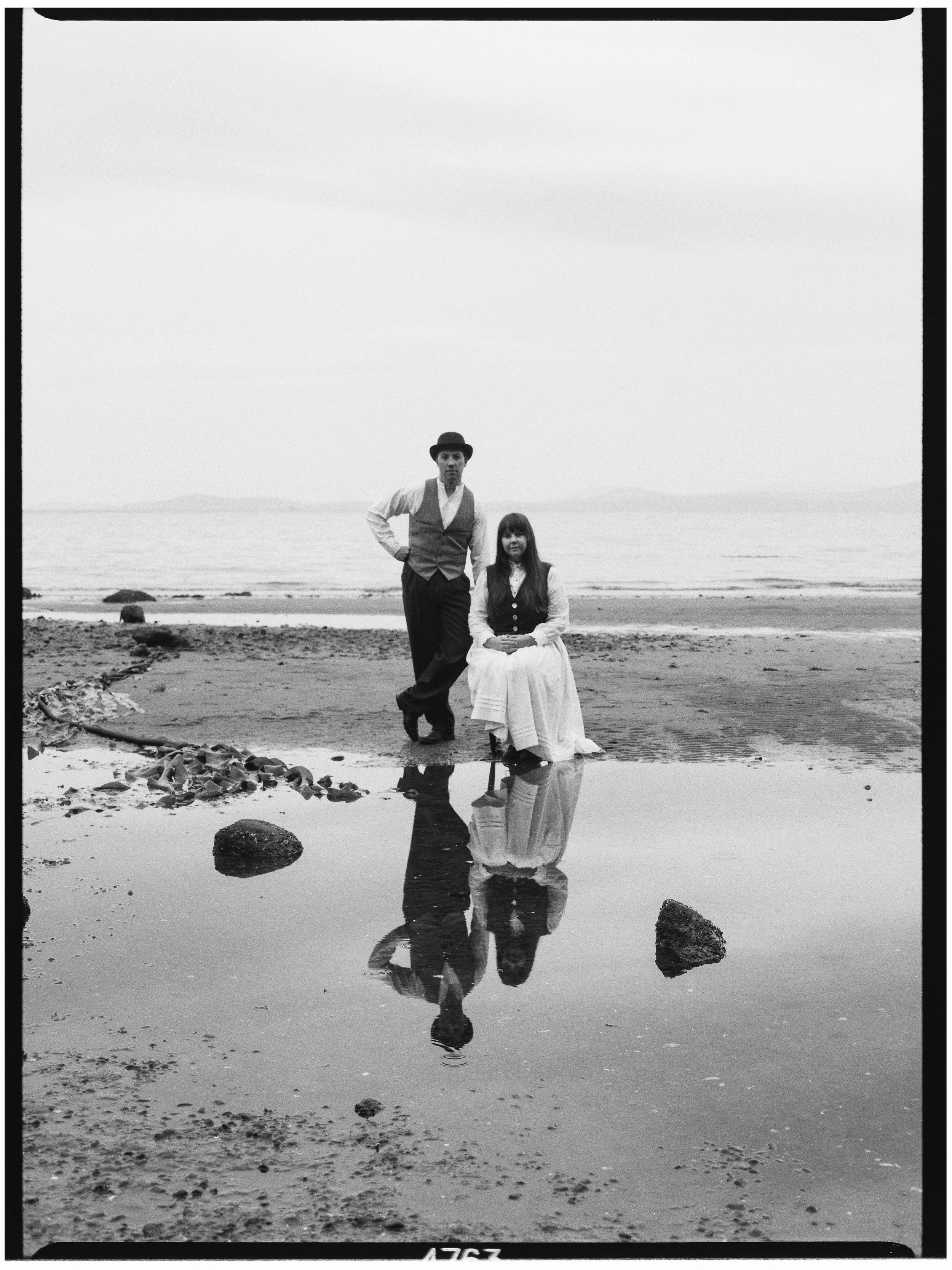 A black and white photo of a man and woman at the beach. The woman is sitting on the sand, and the man is standing nearby with his hand on his hip. Their reflection can be seen in a small puddle of water in front of them. The ocean is in the backgrou