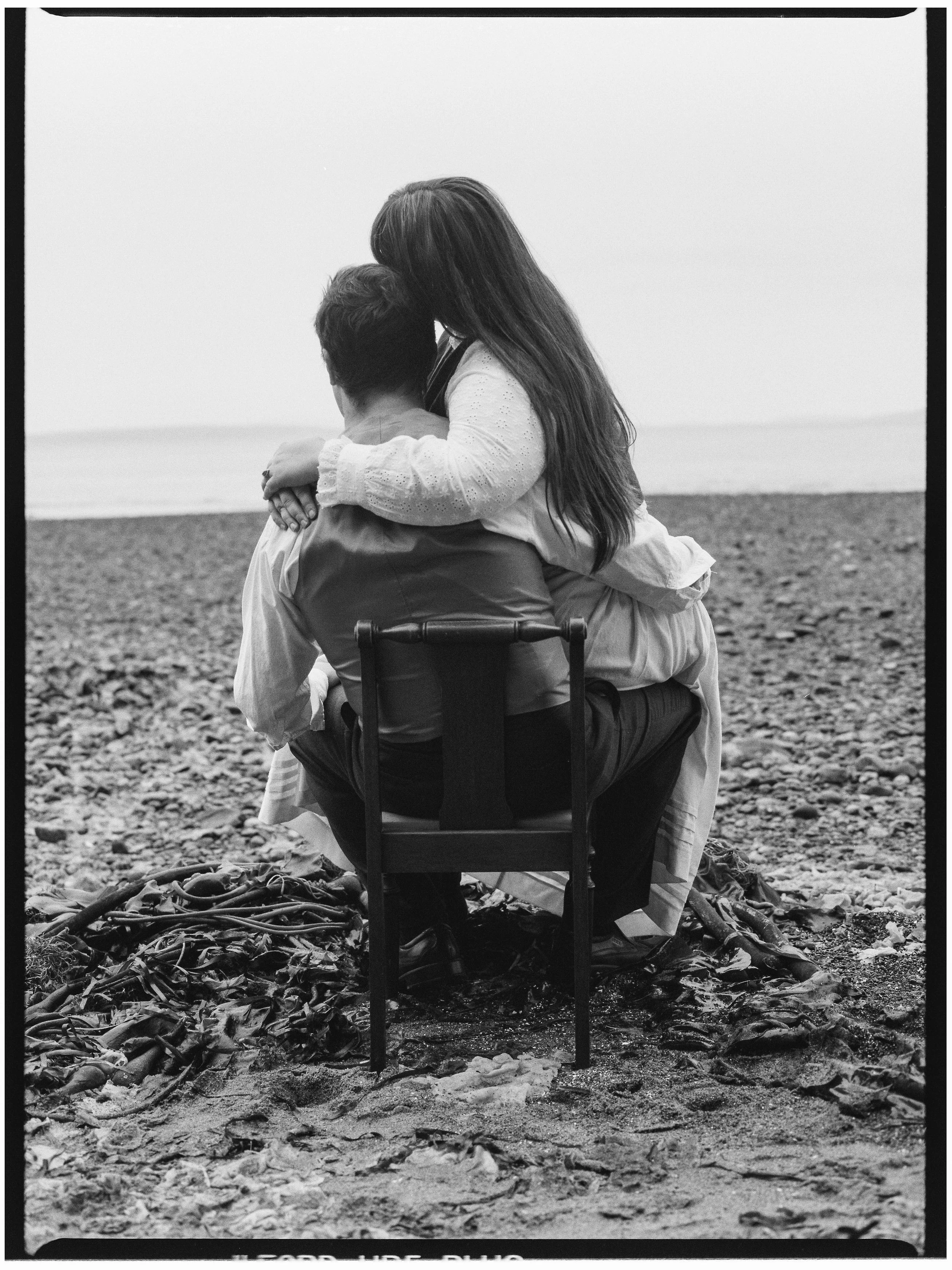 A black and white photo of a woman and a man embracing on a pebble beach, with the woman sitting on the man's lap and holding him close, overlooking the ocean.