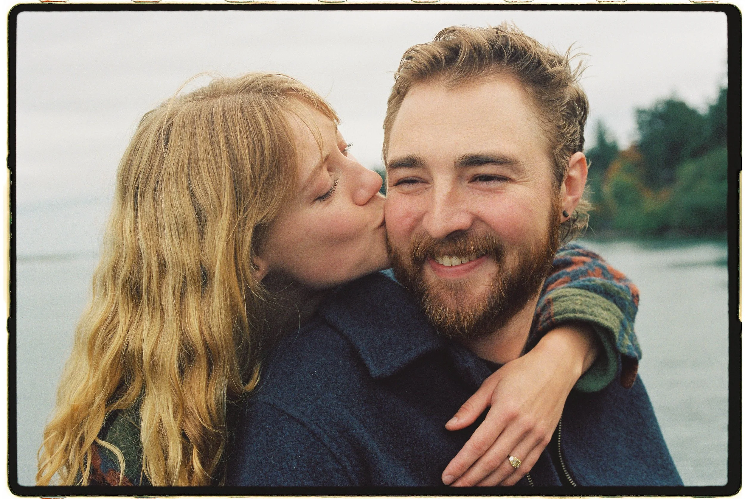A woman with red hair is kissing a man's cheek while hugging him from behind. The man has a beard and is smiling. They are outdoors near a body of water with trees in the background.
