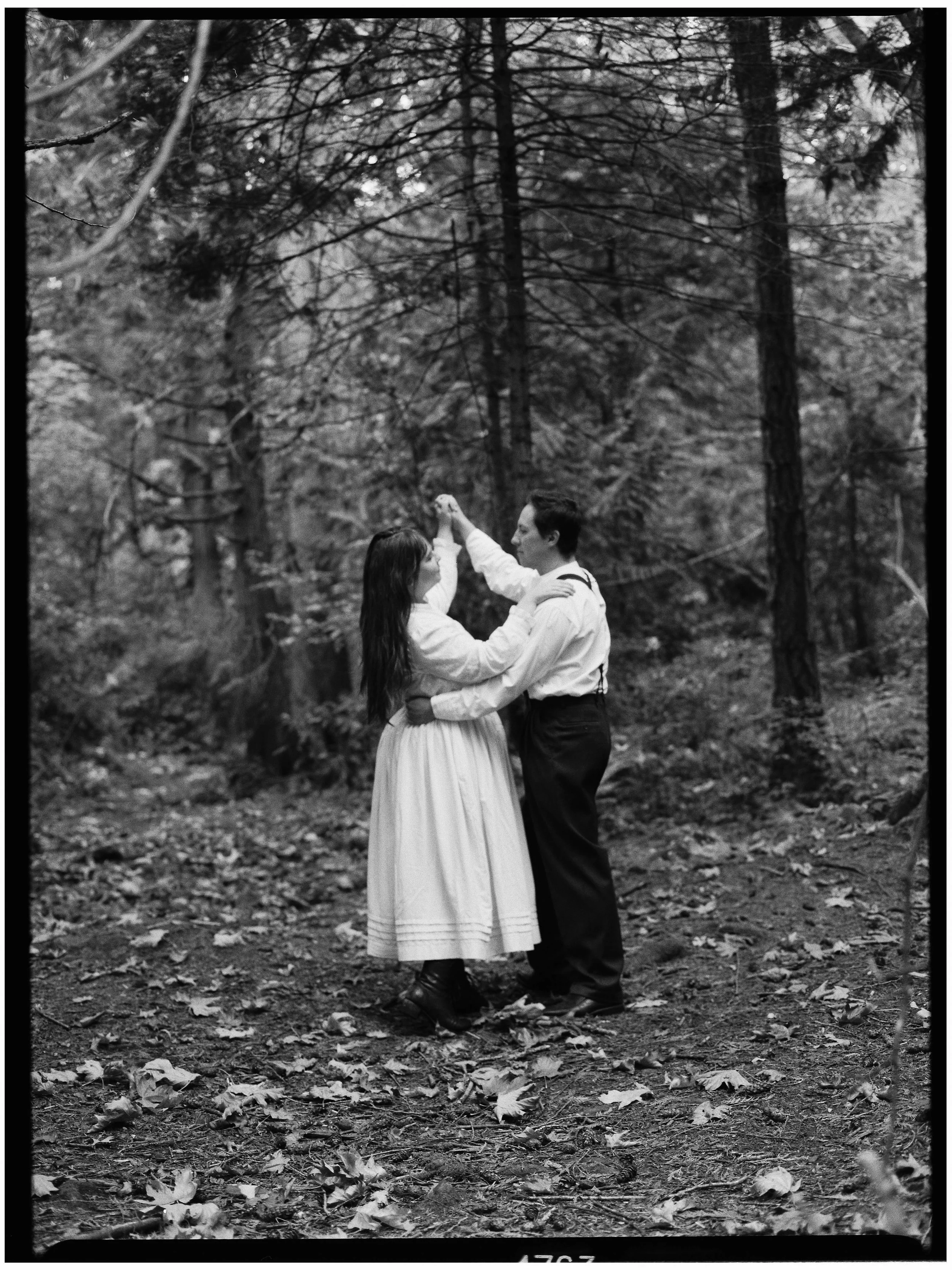 A black and white photo of a couple dancing in a wooded outdoor area with fallen leaves on the ground.
