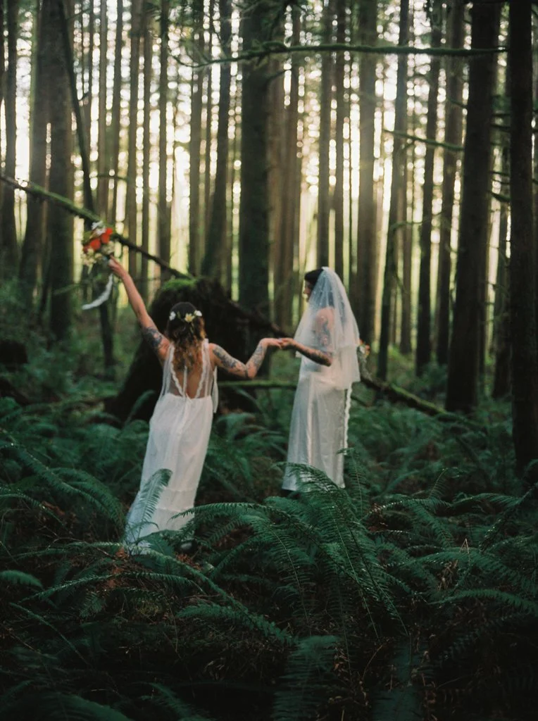 Two women in wedding dresses holding hands in a lush forest, with one woman holding a bouquet and the sun shining through the trees.
