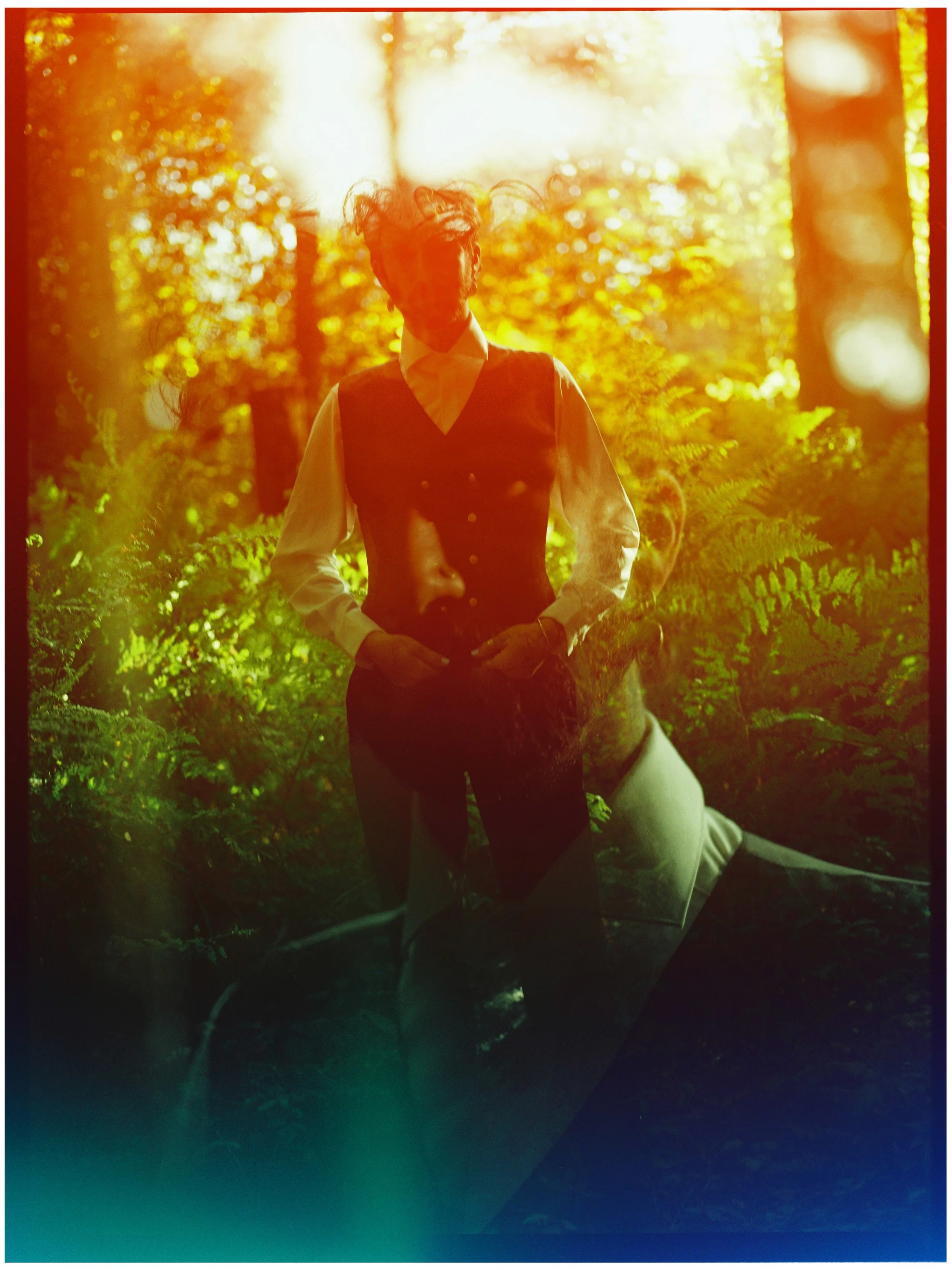 A person in formal attire, wearing a vest and white shirt, standing outdoors in a forest during sunset, with sunlight filtering through trees and illuminating the scene with warm colors.