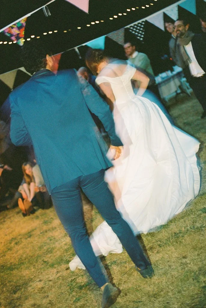 Couple dancing at a wedding reception, with the bride in a white gown and the groom in a blue suit, surrounded by other guests.