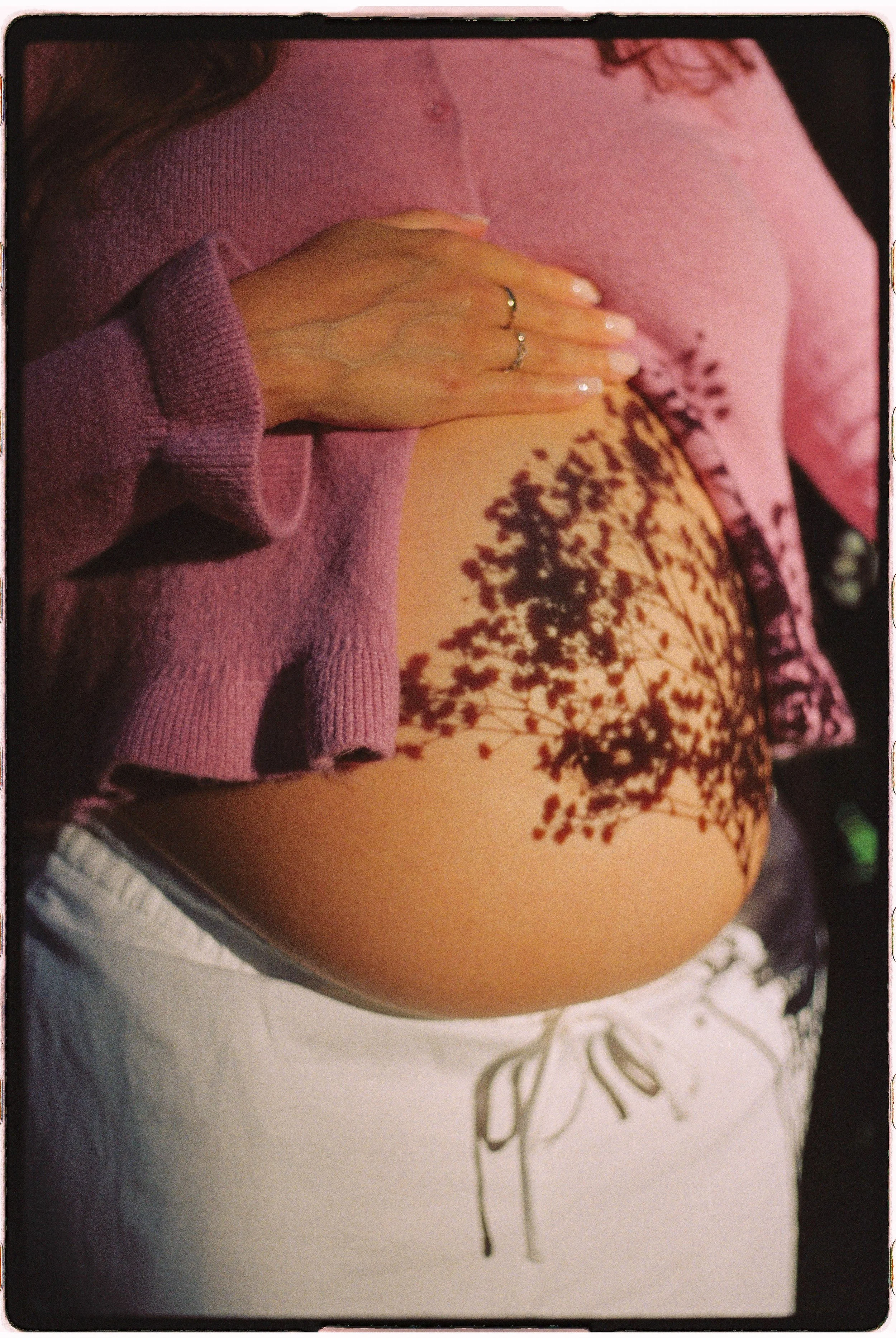 A woman with a tattoo of tree branches on her abdomen, wearing a pink sweater and white pants, resting her hand on her belly with rings on her fingers.