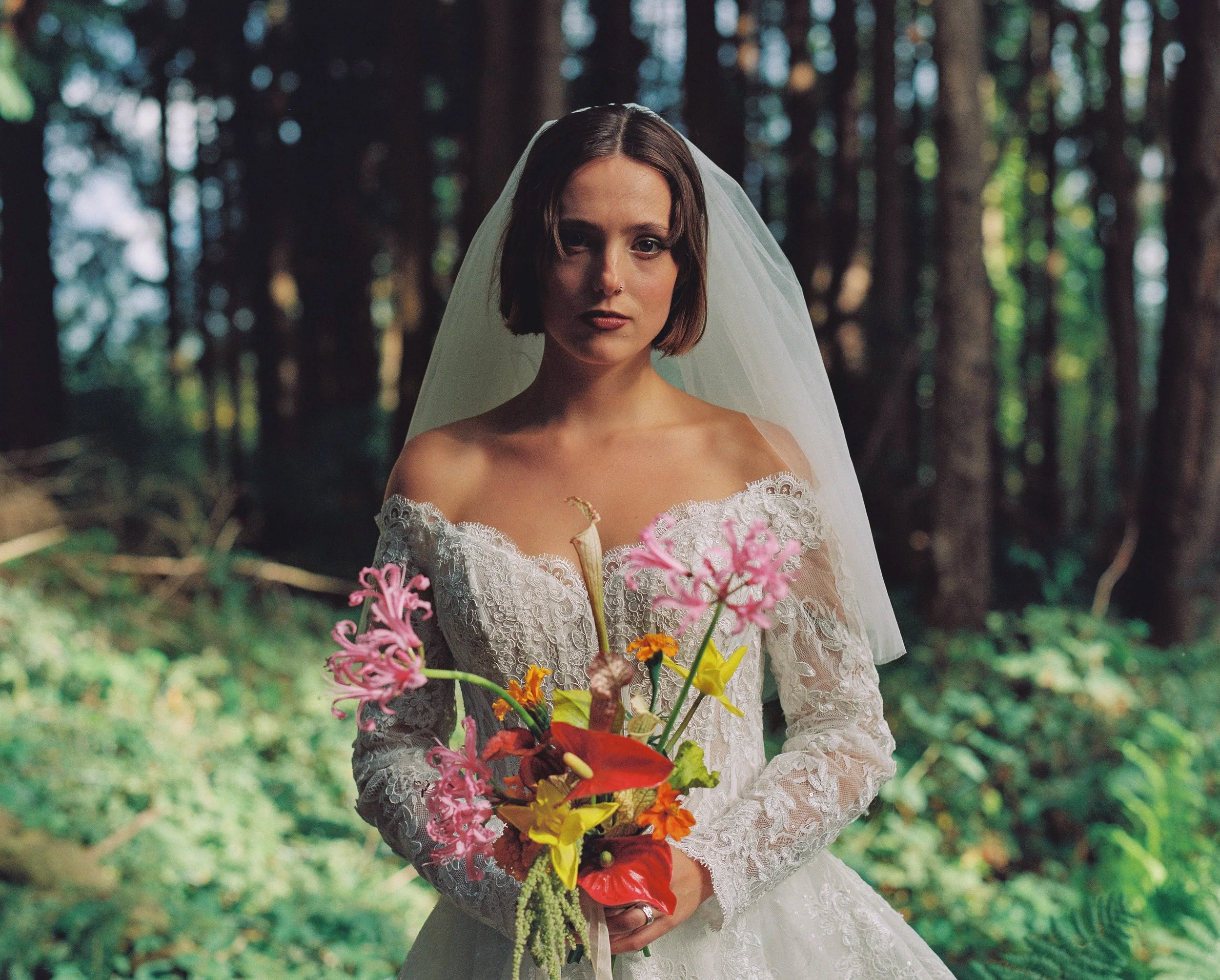 A woman in a white wedding dress and veil holding a colorful bouquet of flowers standing in a forest.