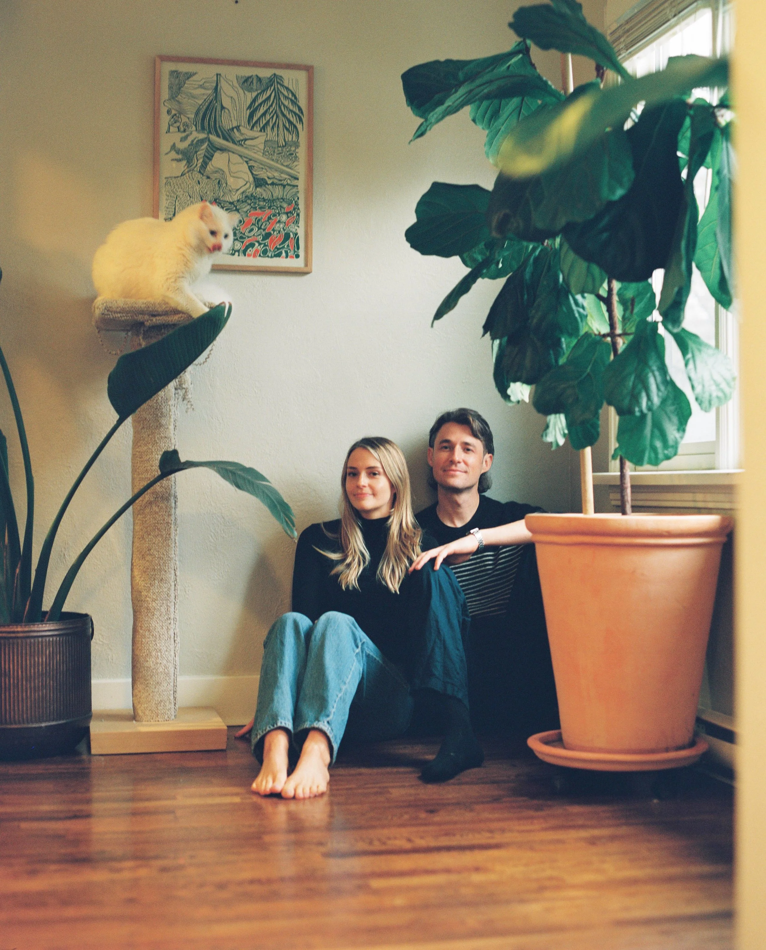 A young woman and man sitting on a wooden floor, surrounded by large houseplants, with a cat on a cat tree to their left and a framed artwork on the wall behind them.