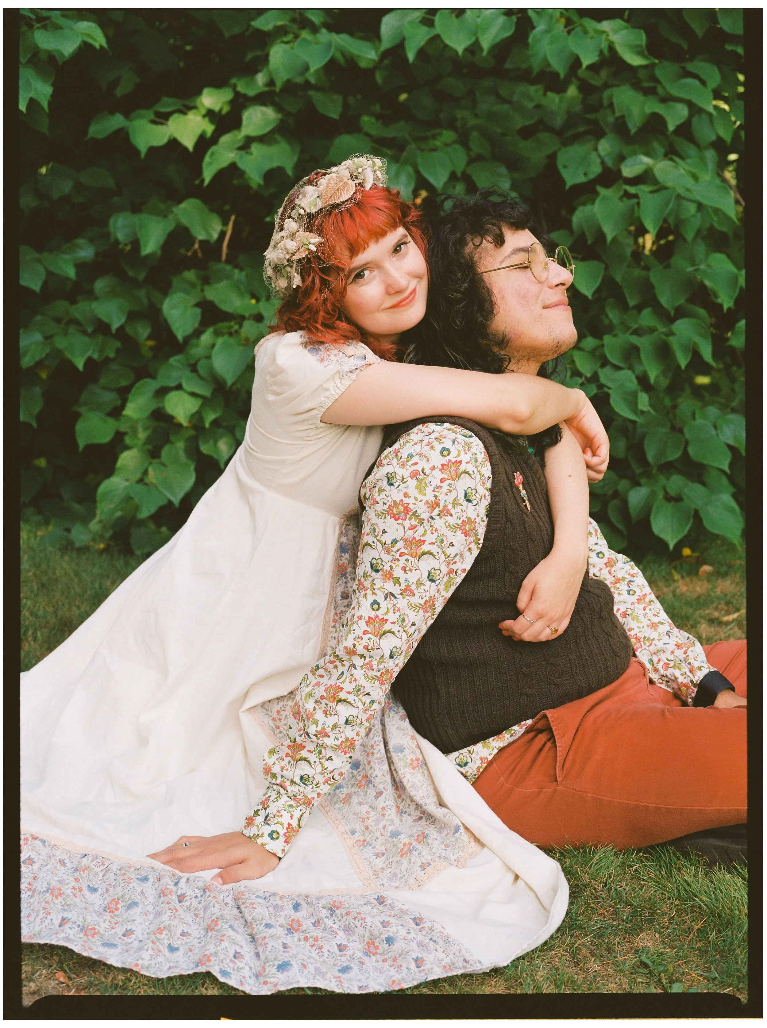 A young woman with curly red hair and a floral headband hugs a man with black curly hair and glasses, sitting on the grass in front of green leafy plants.