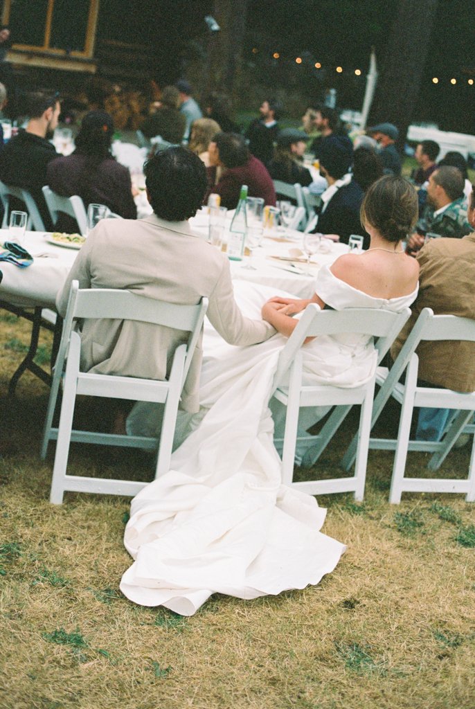 A couple dressed in wedding attire holding hands at a dinner gathering outdoors during nighttime, with other guests seated at tables in the background.