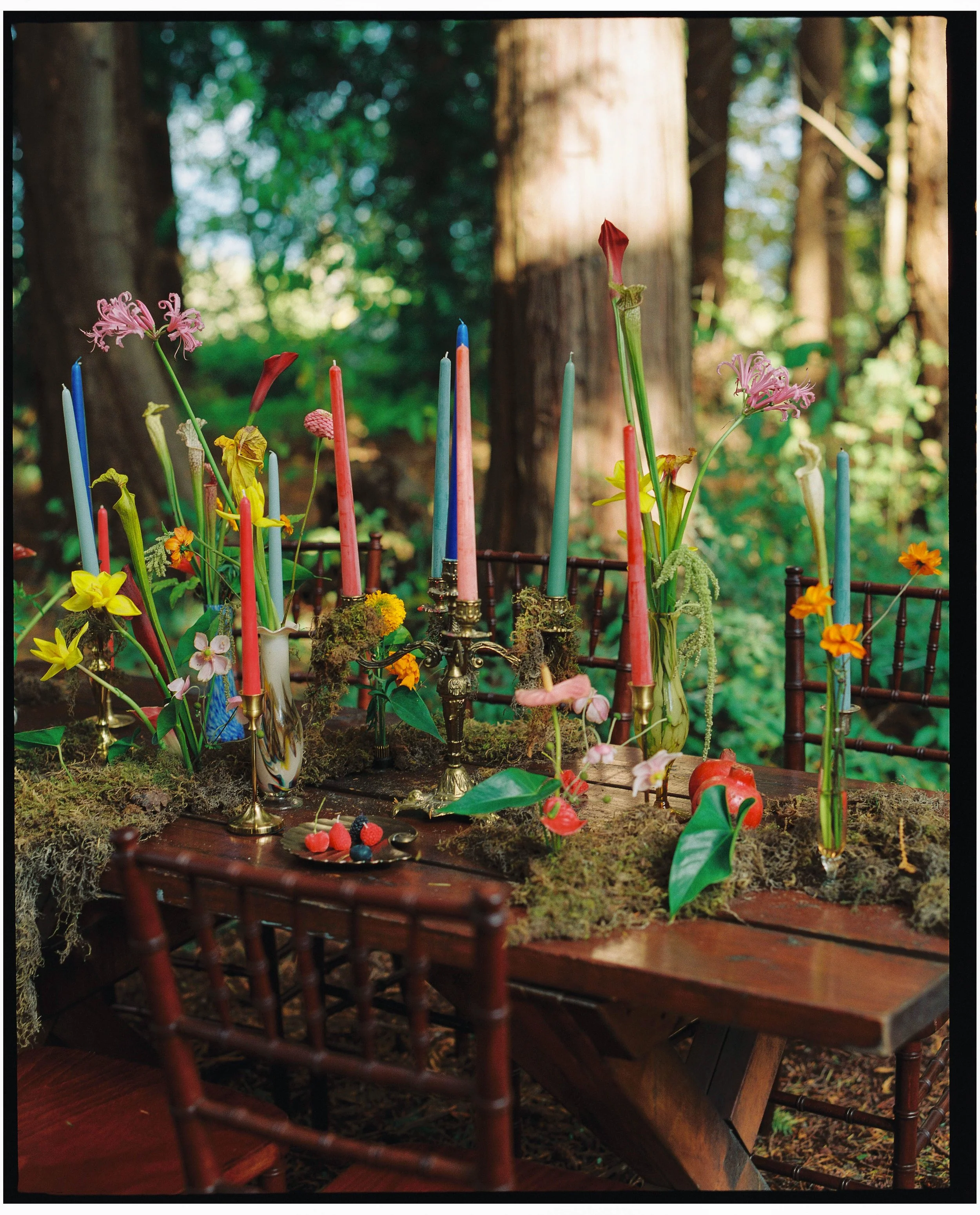 A wooden table in an outdoor forest setting decorated with colorful tall candles in vases, flowers, and moss. The scene appears whimsical and natural with trees in the background.