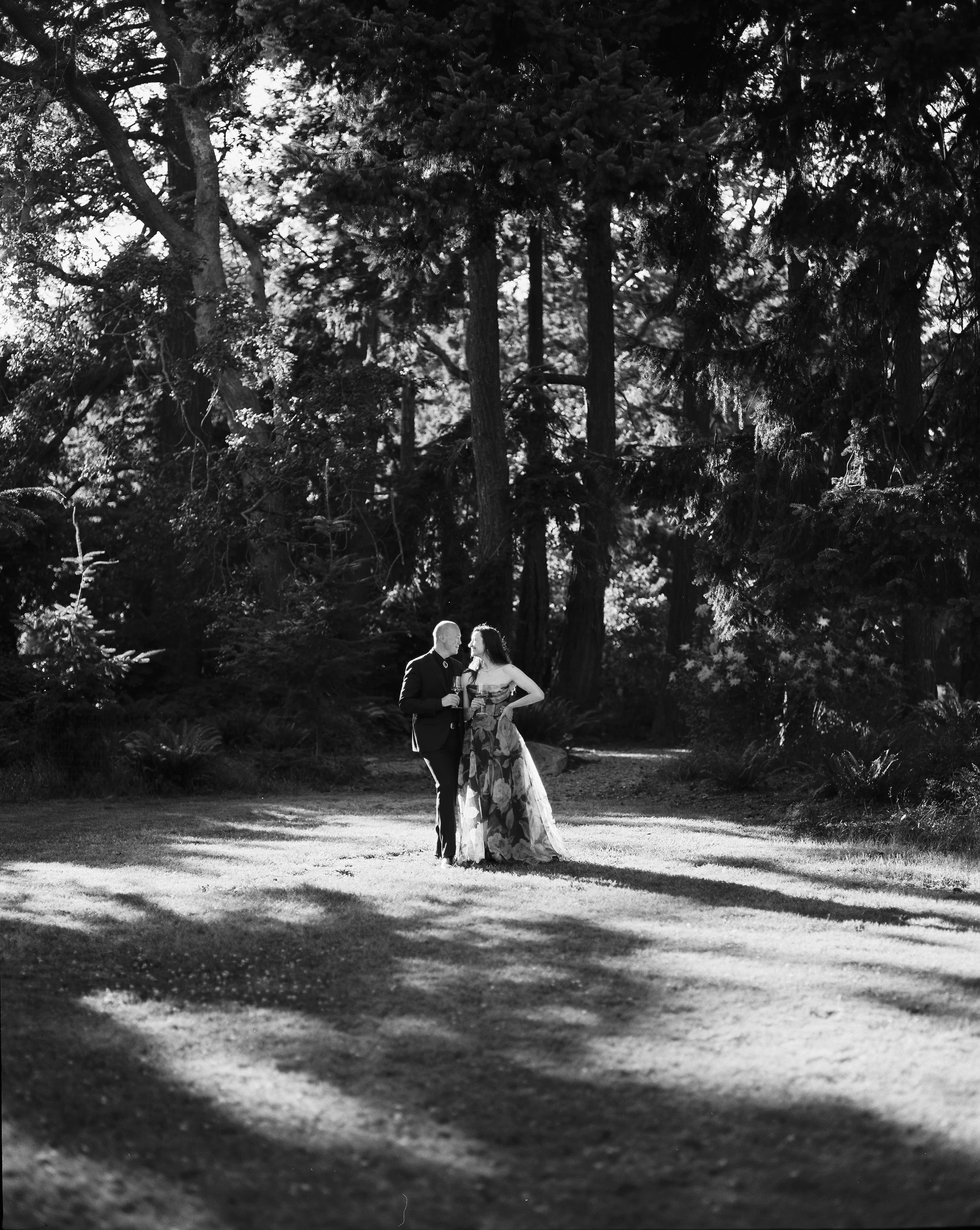 A black-and-white photo of a couple standing on a grassy area among tall trees, looking at each other, with sunlight filtering through the trees.