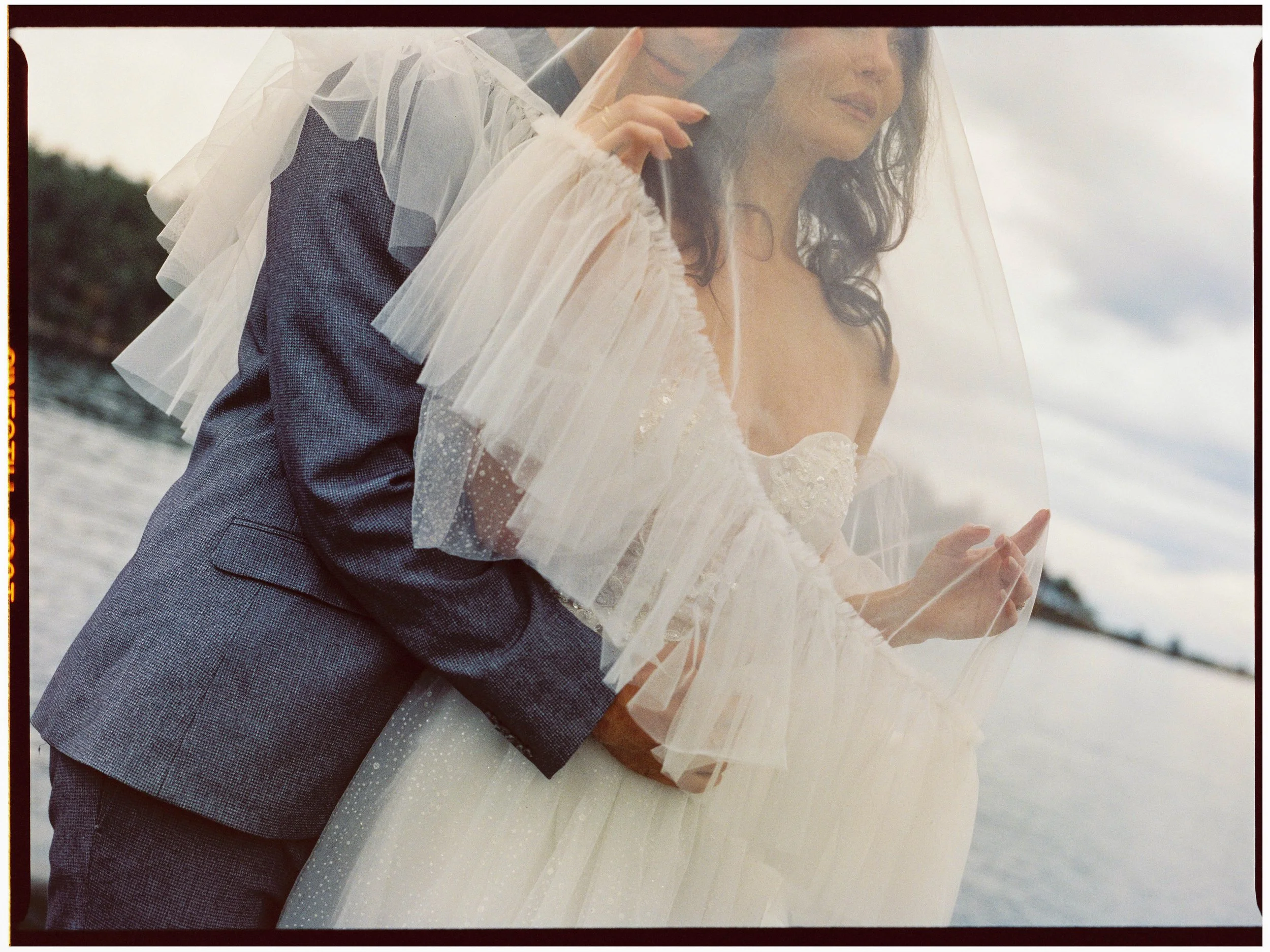 Wedding couple embracing by the water with the bride wearing a white dress and veil, and the groom in a dark suit.
