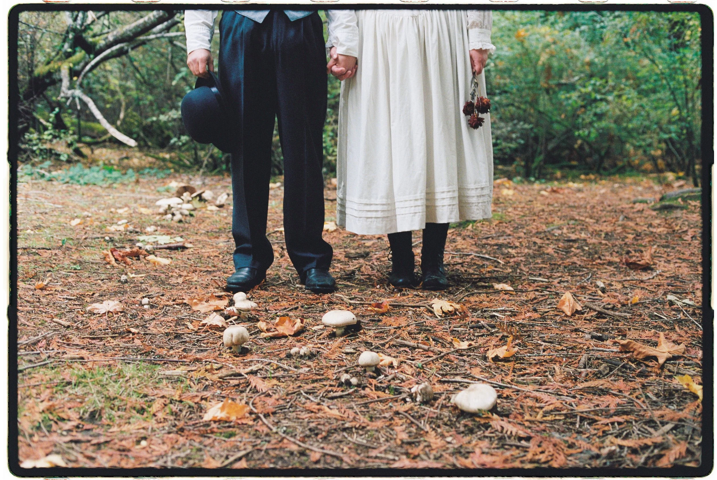 A couple standing in a forest holding hands, with mushrooms on the ground and fallen leaves around them.