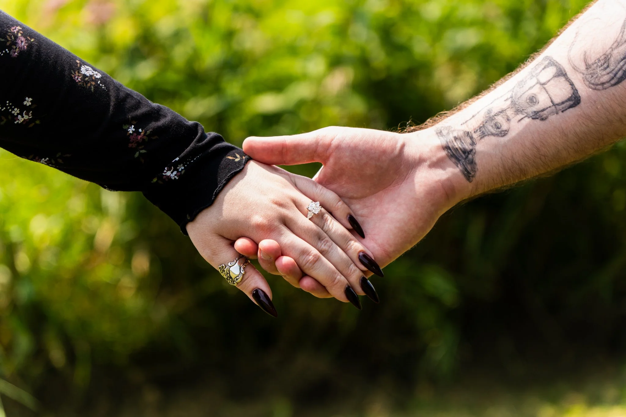 Two people holding hands, one wearing a black long sleeve shirt with floral pattern, the other with tattoos on their arm, and showing an engagement ring.