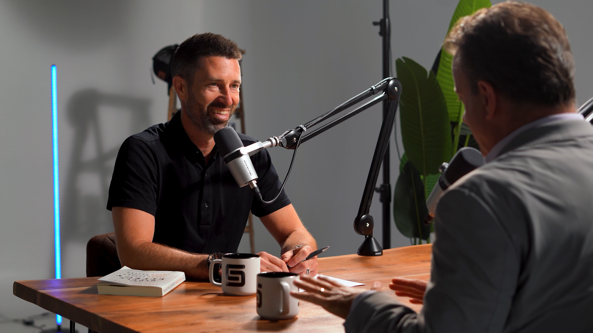 Two men having a podcast recording session at a wooden table with microphones, mugs, and a book in a studio with a large green leaf plant in the background.