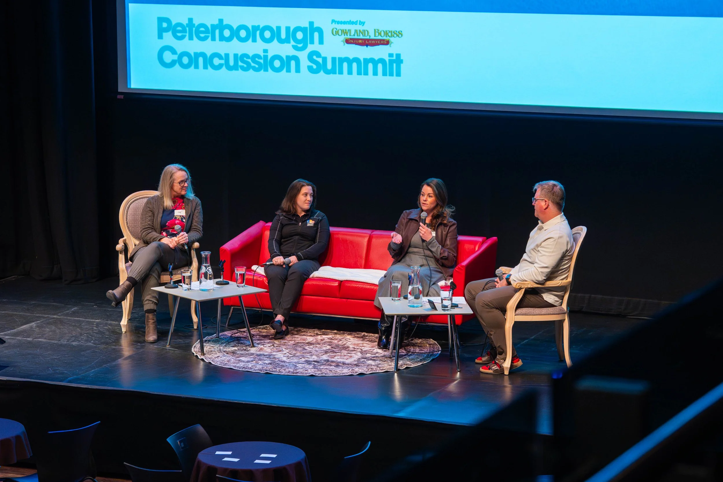 A panel discussion at the Peterborough Concussion Summit featuring four individuals seated on stage with a large screen displaying the event's title behind them.