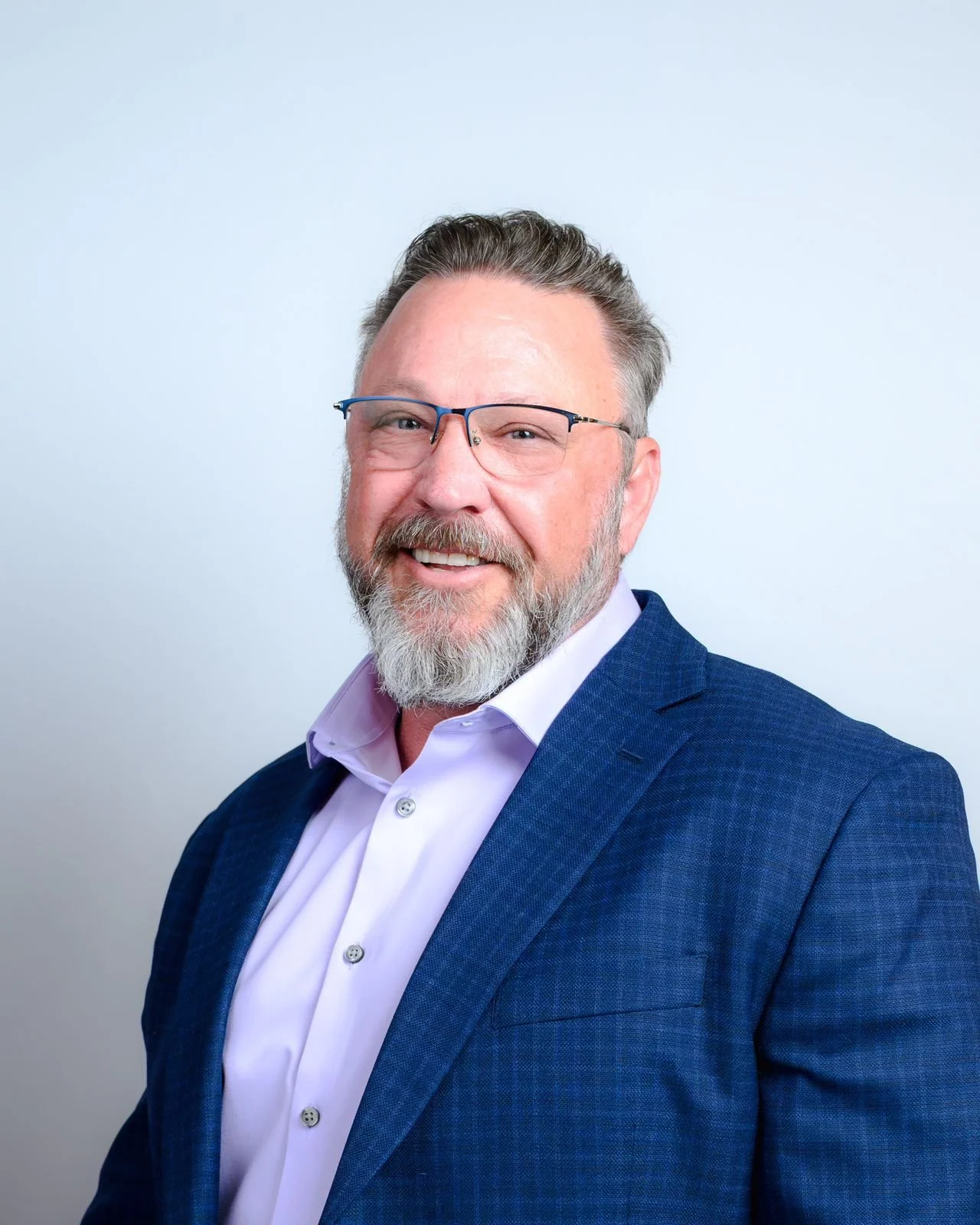 Professional man with glasses, gray beard, and short gray hair, smiling, wearing a blue suit jacket and light purple shirt, against a plain light background.