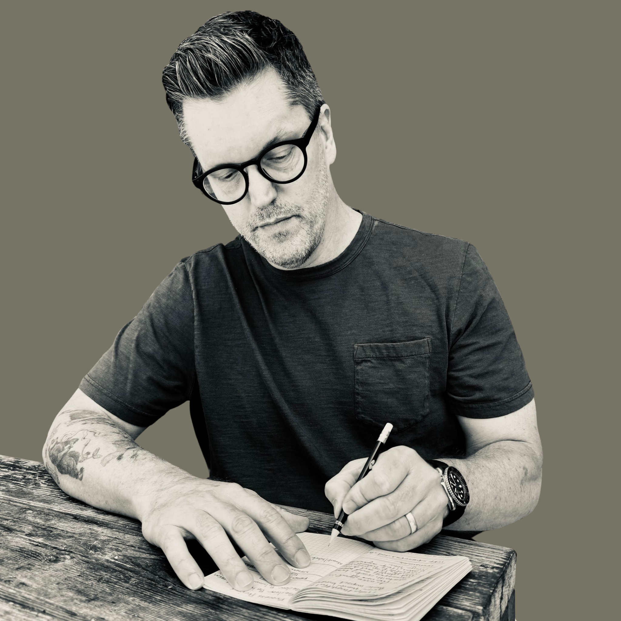 A man with glasses and tattoos on his arm is writing in a notebook with a pen while sitting at a wooden table.