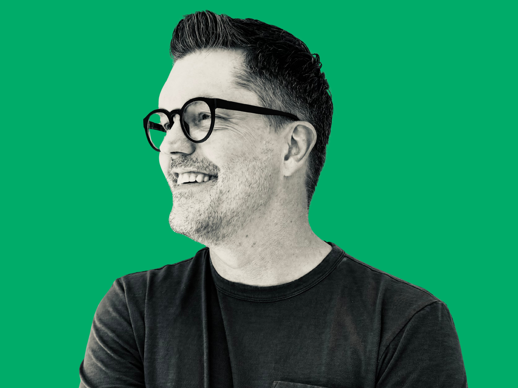 A man with glasses and dark hair, smiling, wearing a black shirt, against a green background.