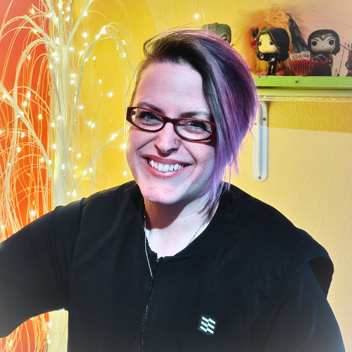 Smiling woman with purple and black hair, glasses, and black jacket, in front of string lights and a yellow wall with shelves holding Funko Pop! figures.