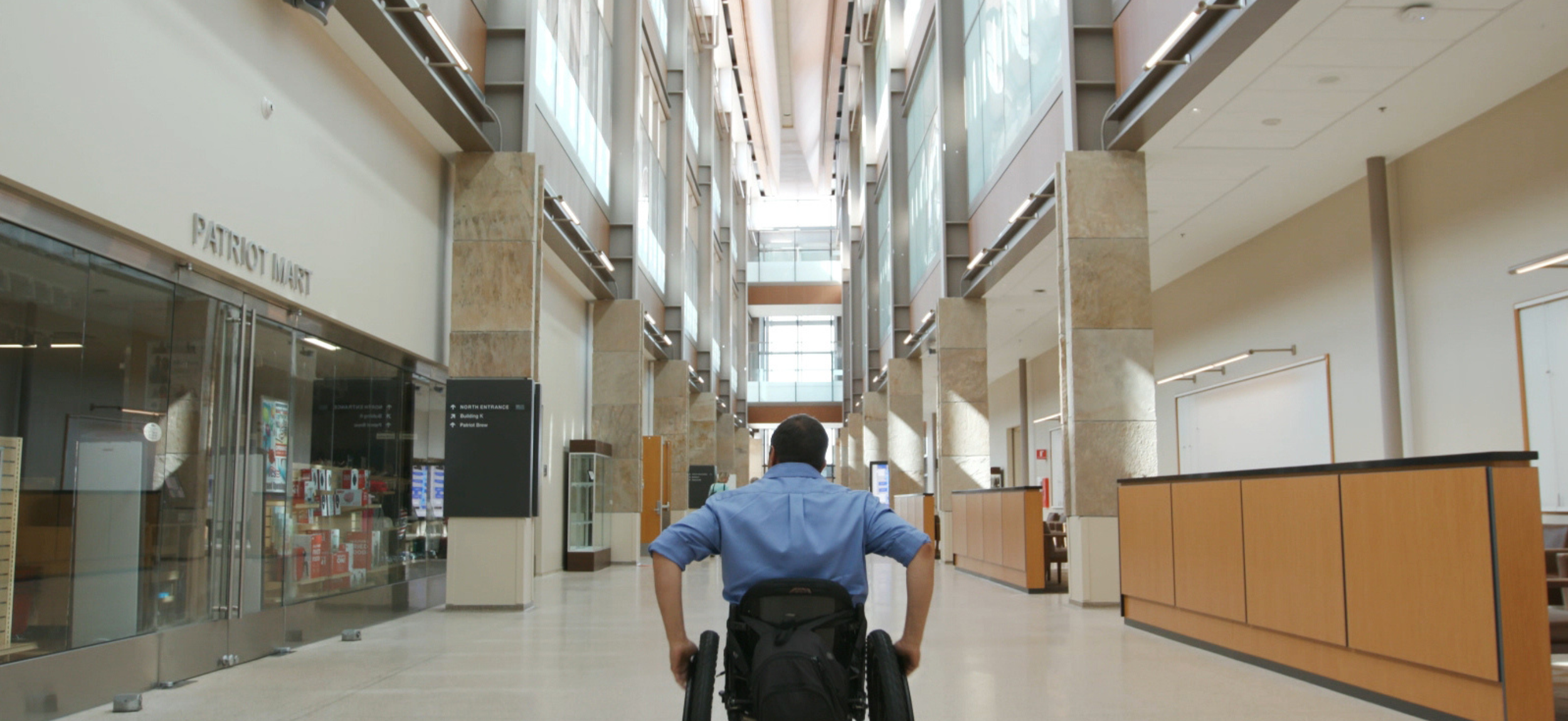 A person in a wheelchair wearing a blue shirt is moving through a spacious, modern indoor public space with high ceilings, large windows, and several levels visible.