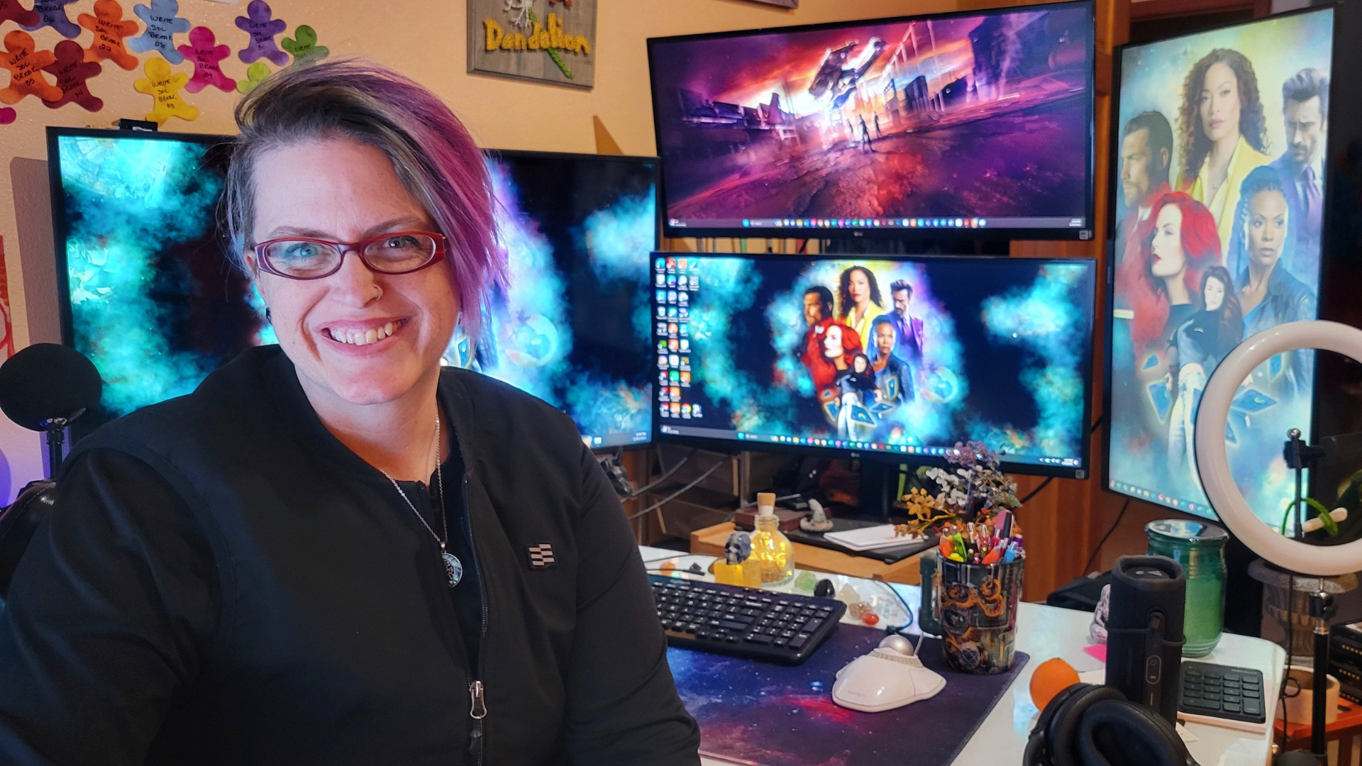 A smiling female content strategist with short hair and glasses sitting in front of multiple computer monitors displaying colorful space-themed and fantasy-themed wallpapers. The workspace includes a ring light, a keyboard, a mouse, a microphone.