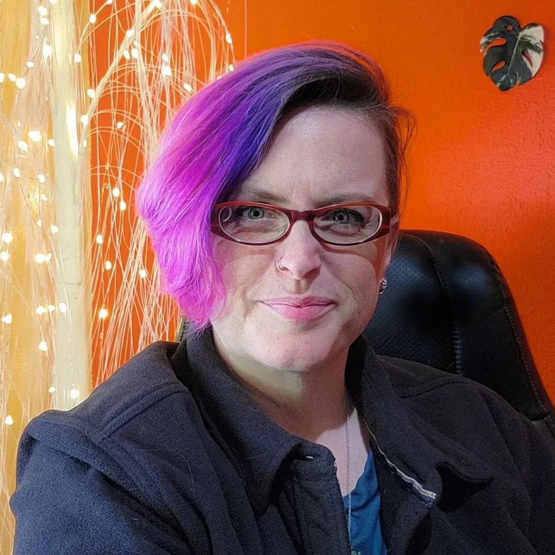 A female content strategist with colorful purple and pink hair, glasses, and a nose ring sitting in front of an orange wall with decorative lights and a black monstera leaf wall hanging.