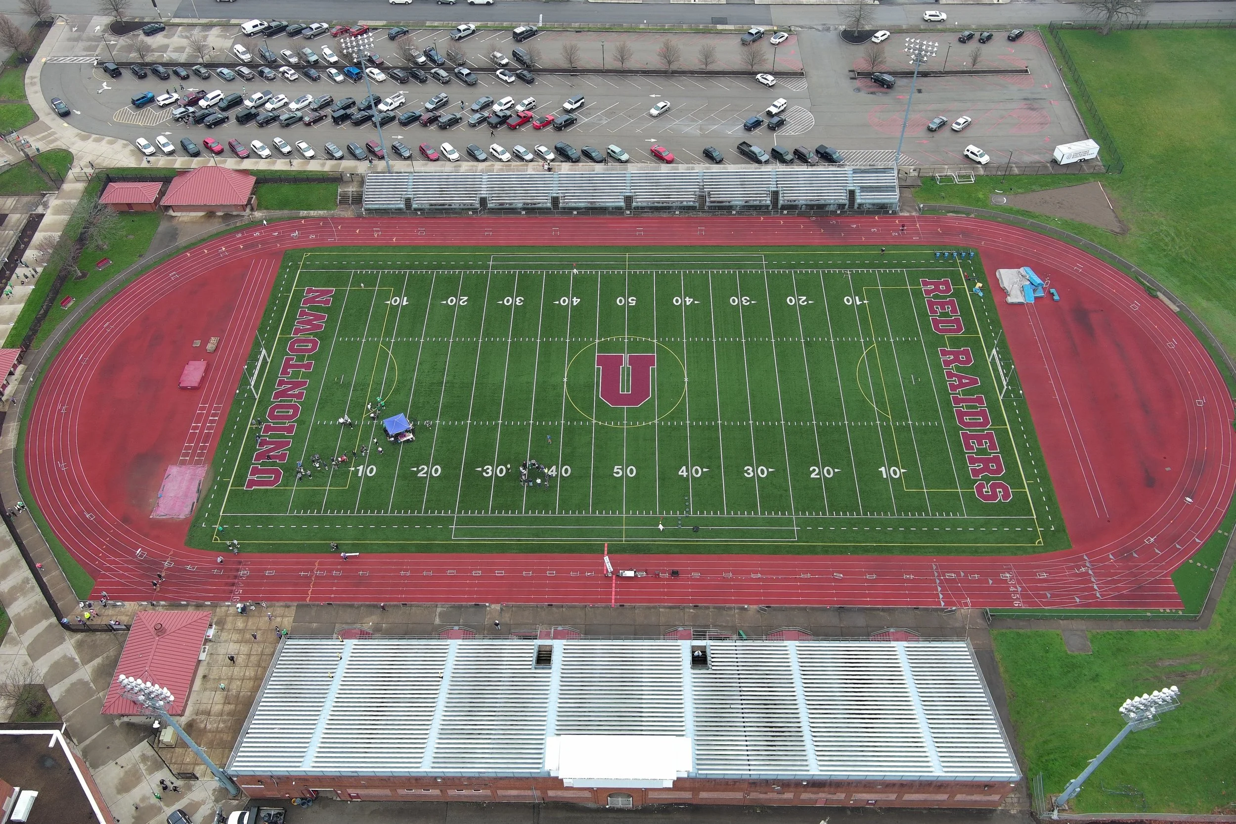 An aerial view of a football field with red running track, labeled 'UNION-MON' on one side and 'RED RAIDERS' on the other, surrounded by parking lot and athletic facilities.