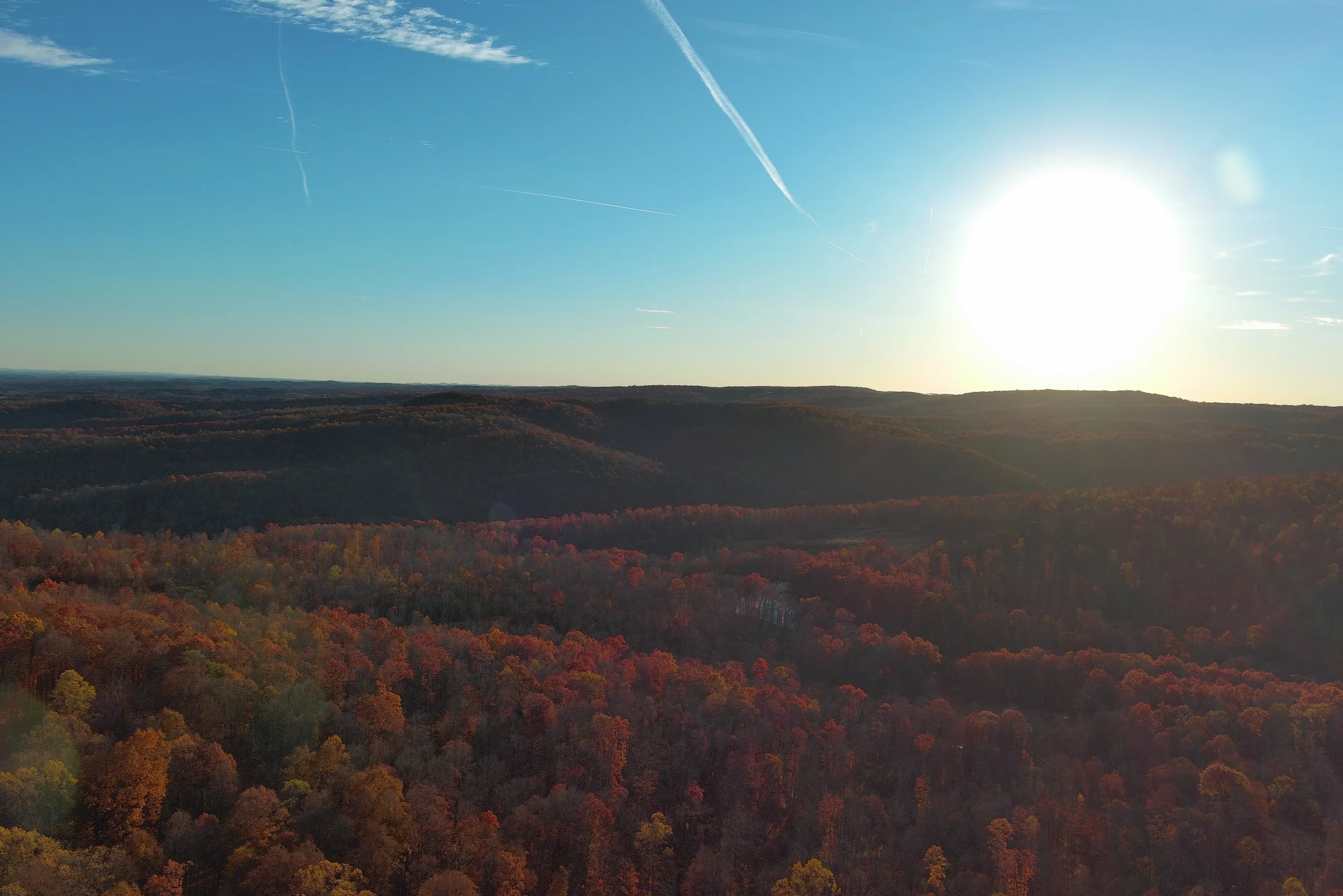 A scenic view of a forested landscape with fall foliage, rolling hills, and a bright sun in the sky during clear weather.