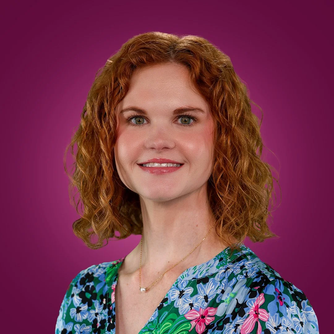 A woman with red curly hair, wearing a floral blouse and a gold necklace with a pearl, posing against a purple background.