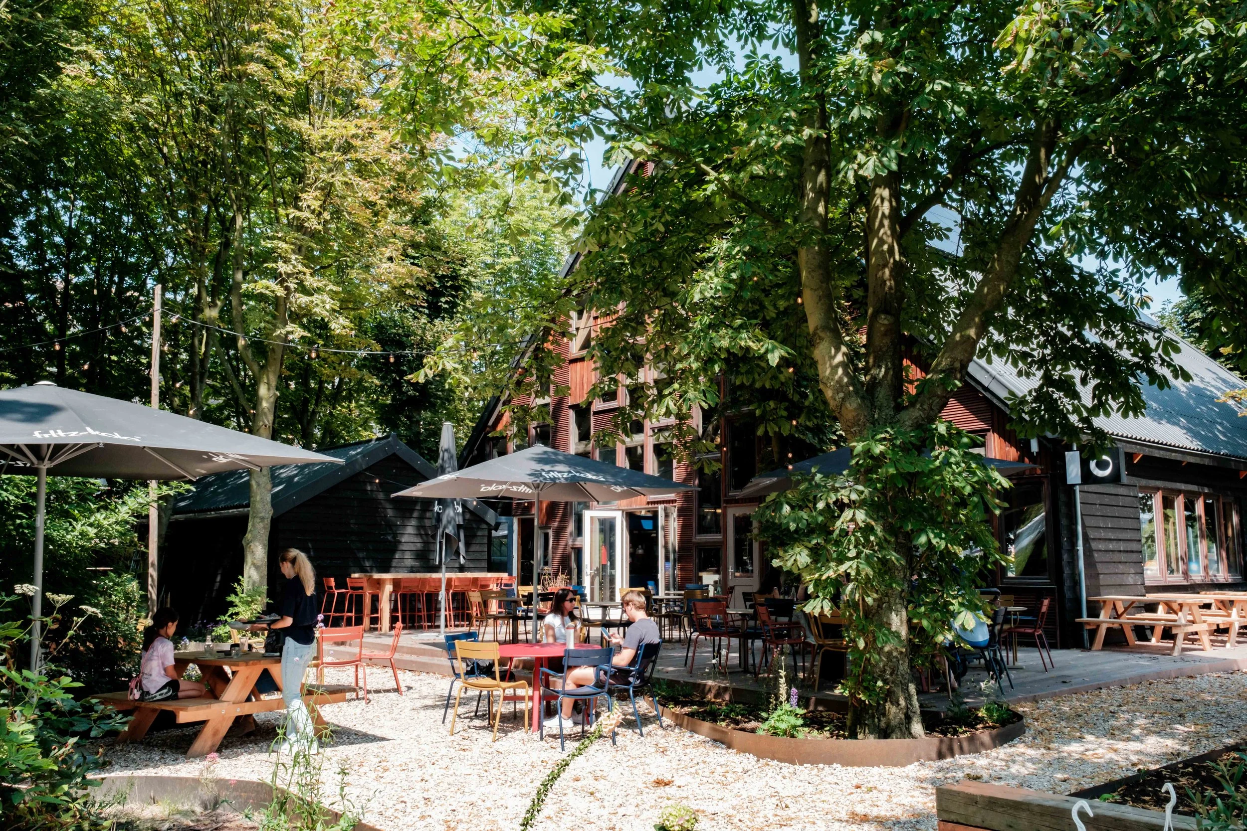 Een buitenterras van een café of restaurant met mensen die zitten te praten en drinken, omgeven door bomen en groen, met een houten gebouw op de achtergrond en parasols voor schaduw.