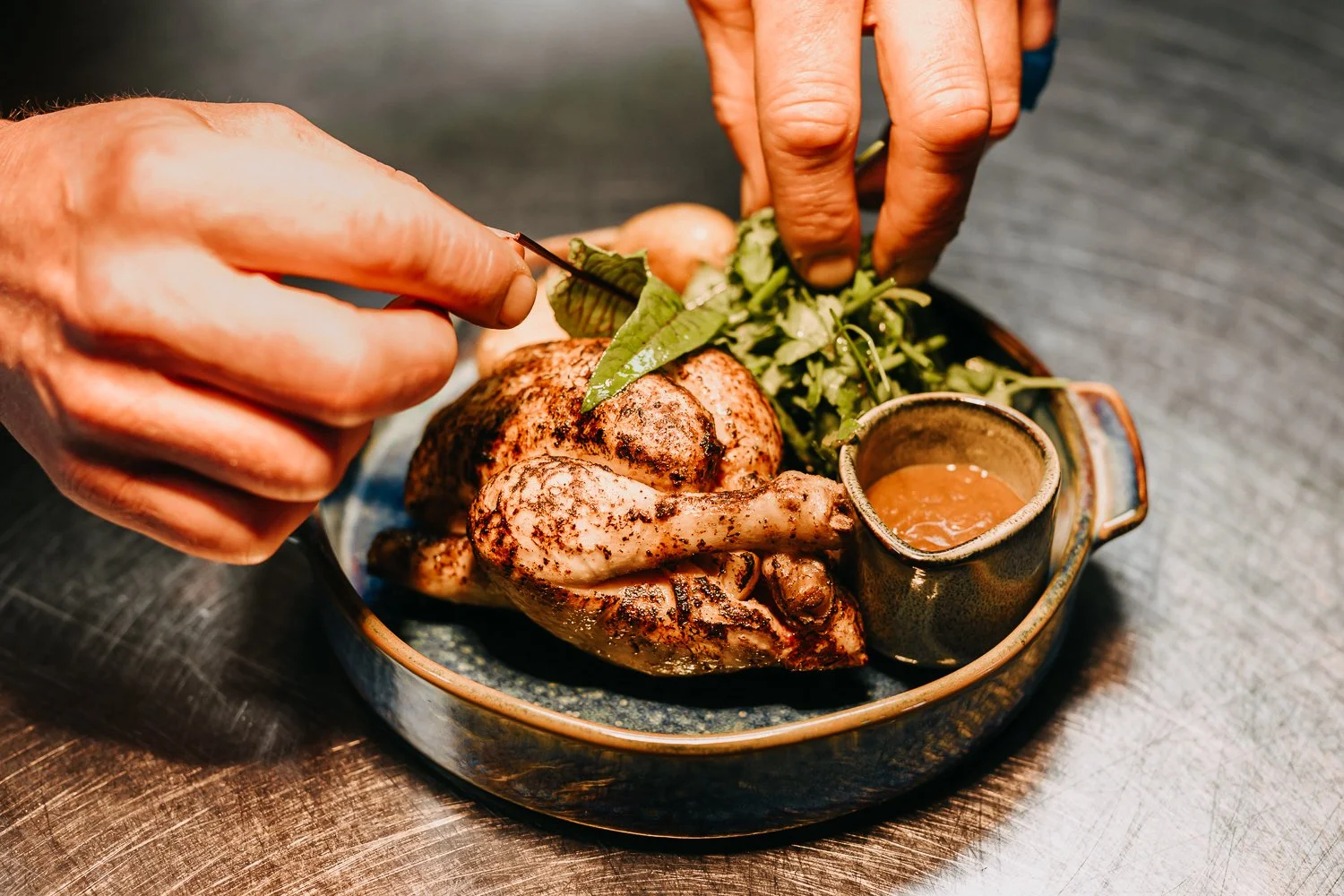 Een geroosterd hele kip op een bord, versierd met groene bladeren en geserveerd met een saus in een klein kopje.