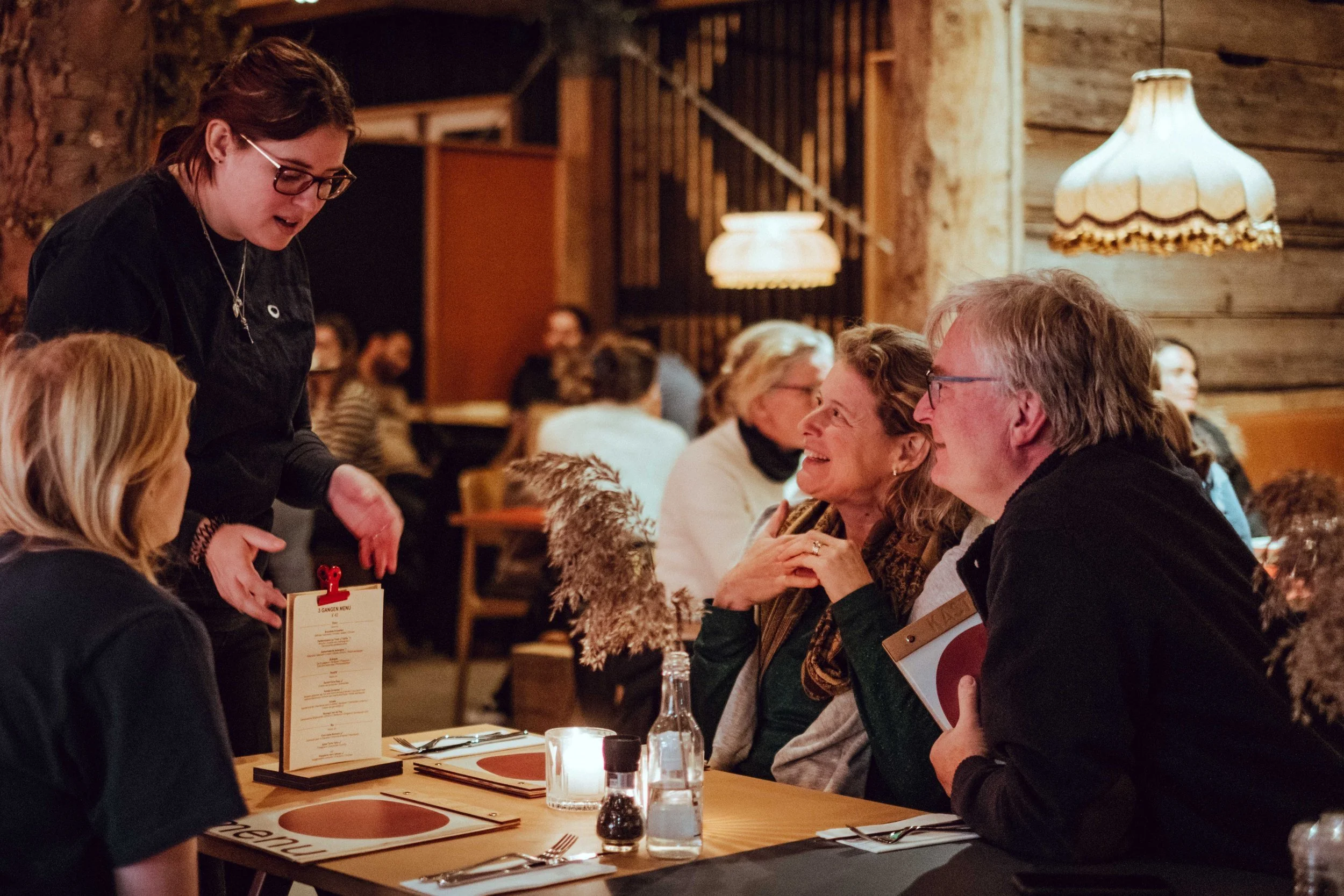 Een gezellige restaurantsetting met drie mensen aan tafel te genieten van een gesprek. Een serveerster staat naast hen en neemt bestellingen op.