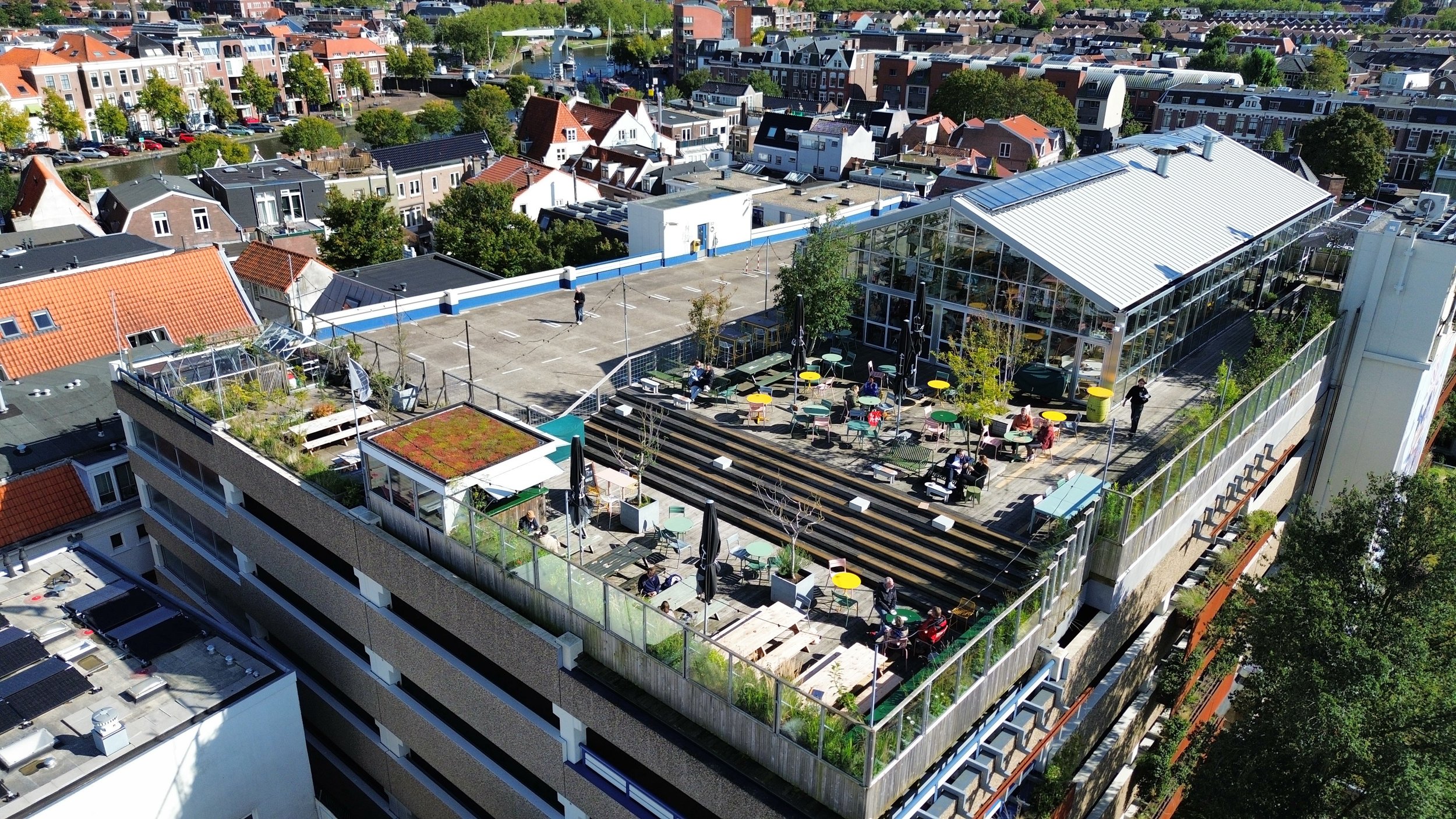 Een hoge gebouwenflat met een terras op het dak, waar mensen zitten en genieten van hun tijd. Het terras heeft kleurrijke tafels en stoelen, en er zijn planten en een kas op het dak.