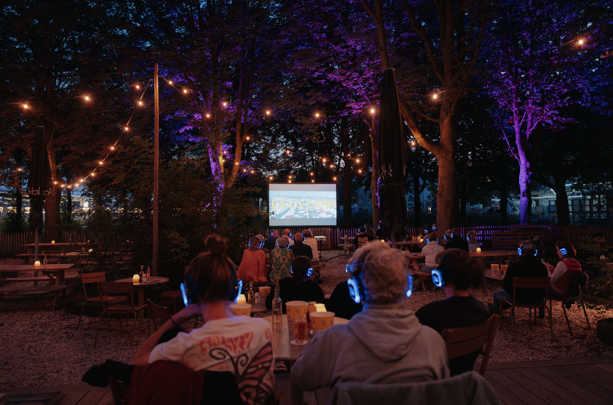 Mensen kijken naar een film onder kleurrijke lichtjes in een bosrijke omgeving.