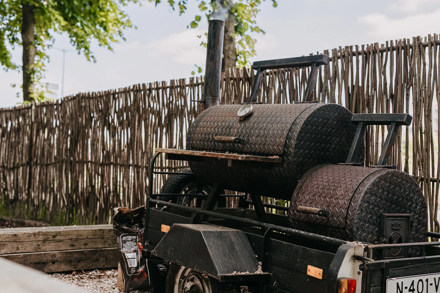 Een oude barbecue op een kleine trailer, geplaatst naast een houten afscheiding en onder een boom. 