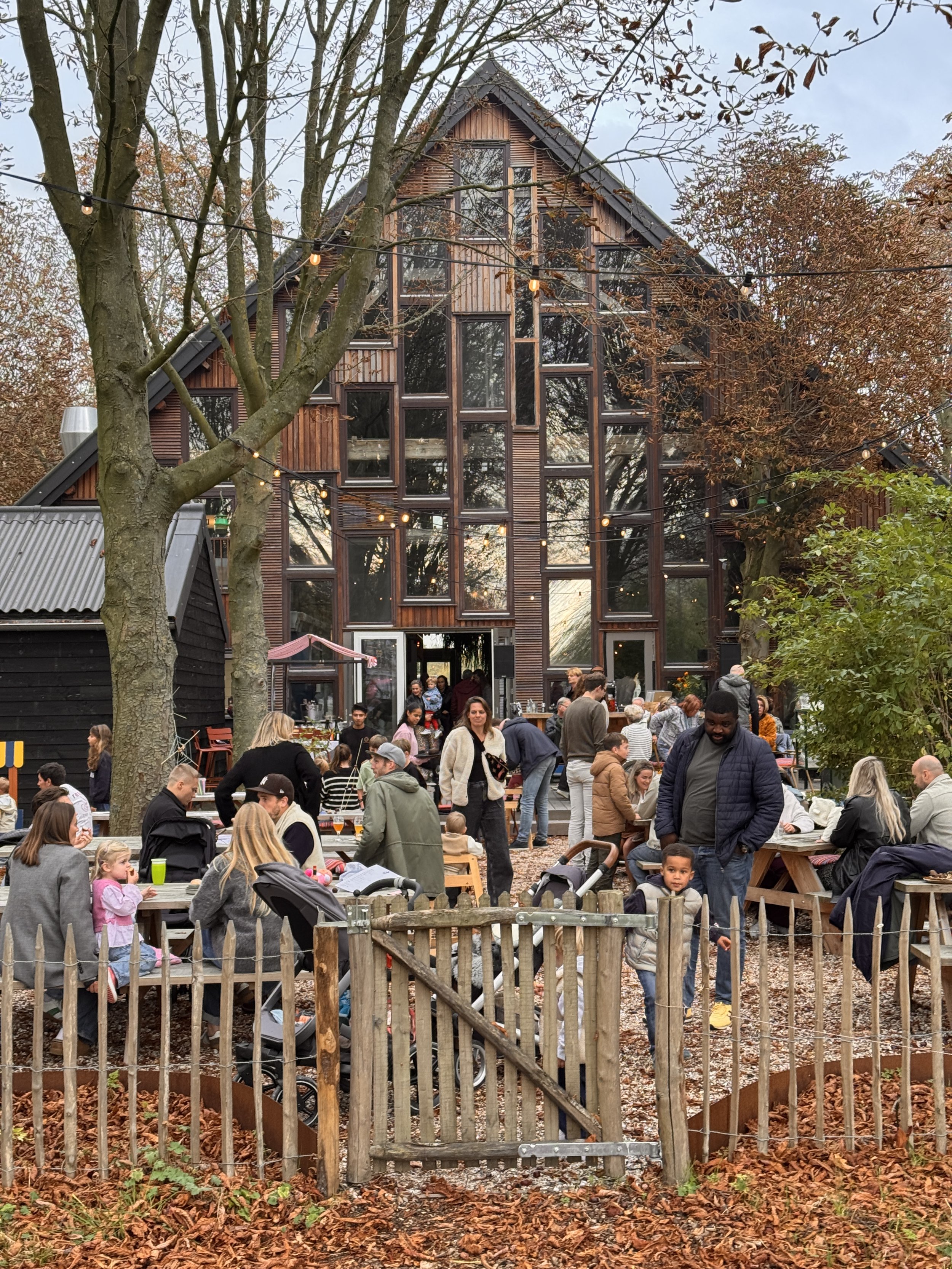 Blije mensen en gezinnen genieten van een buitenfeest in de herfst, met een modern houten huis op de achtergrond en omgeven door bomen met vallende bladeren.
