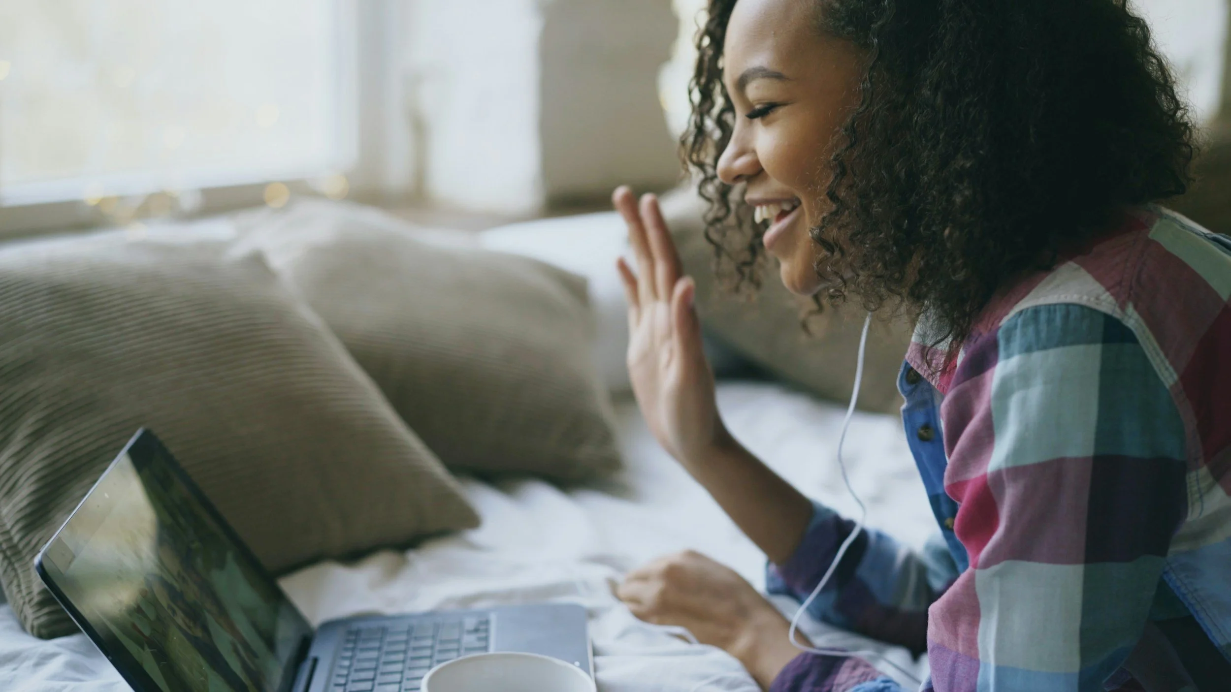 A young woman of color with curly hair smiling and waving while on a video call on her laptop, sitting on a bed with pillows and a mug nearby, in a well-lit room.
