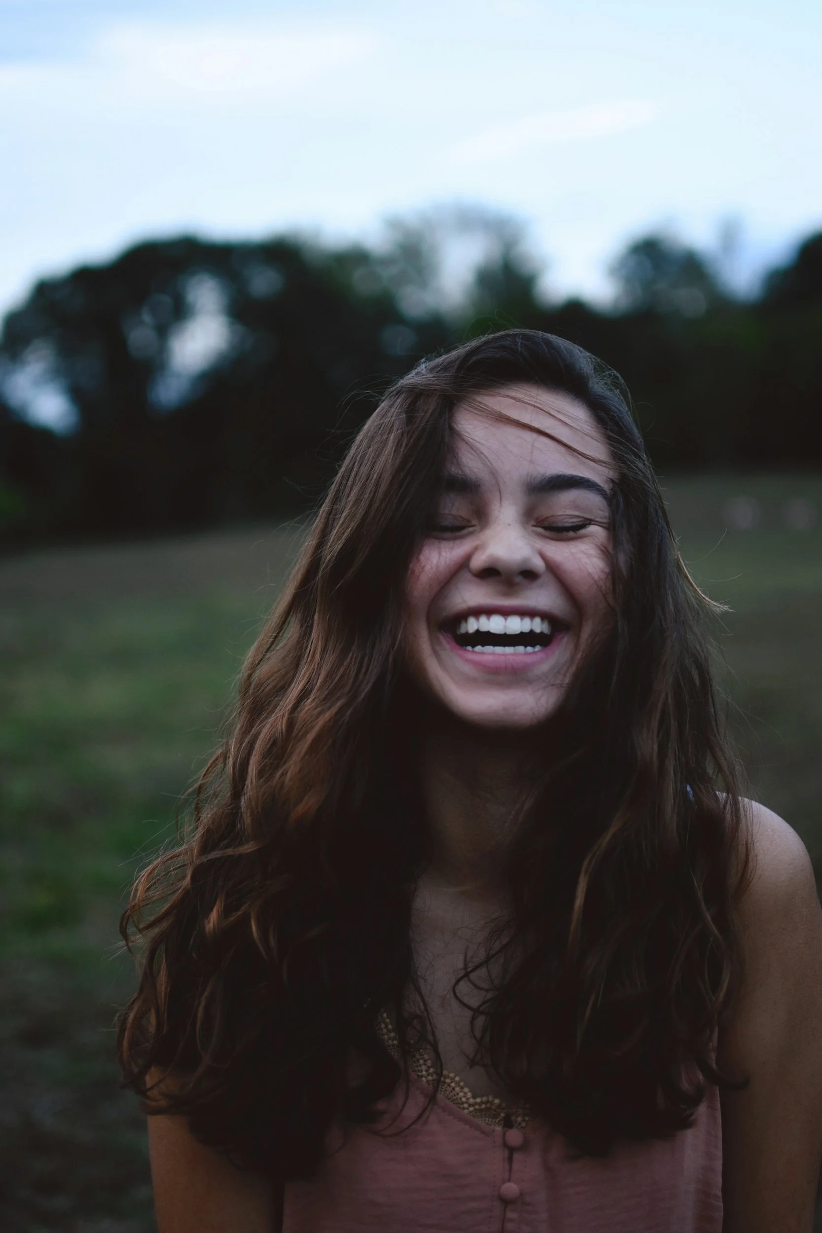 A woman with long, wavy brown hair laughing with her eyes closed, enjoying the outdoors.