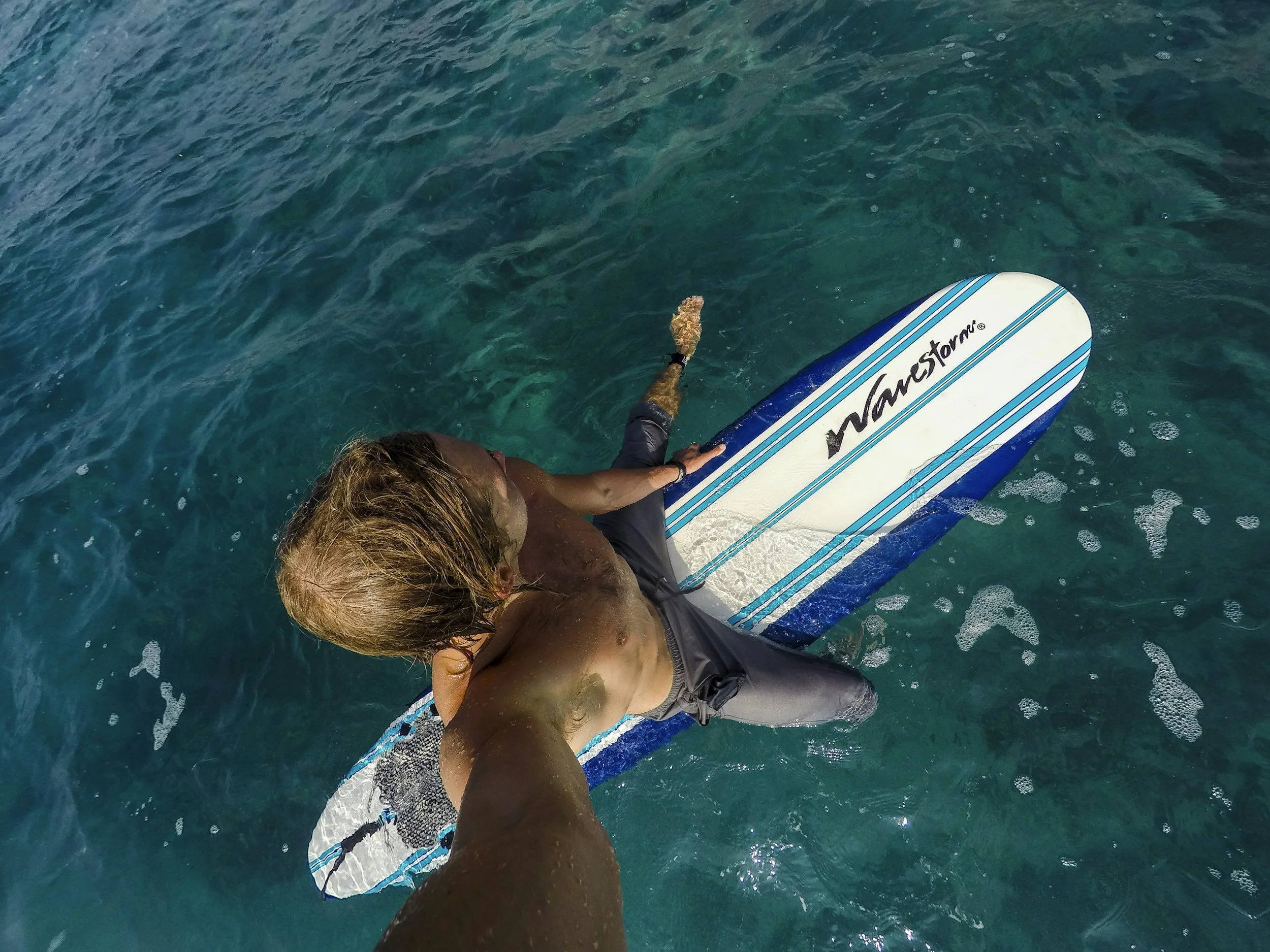 A man is sitting on a blue and white surfboard in calm, clear blue water.