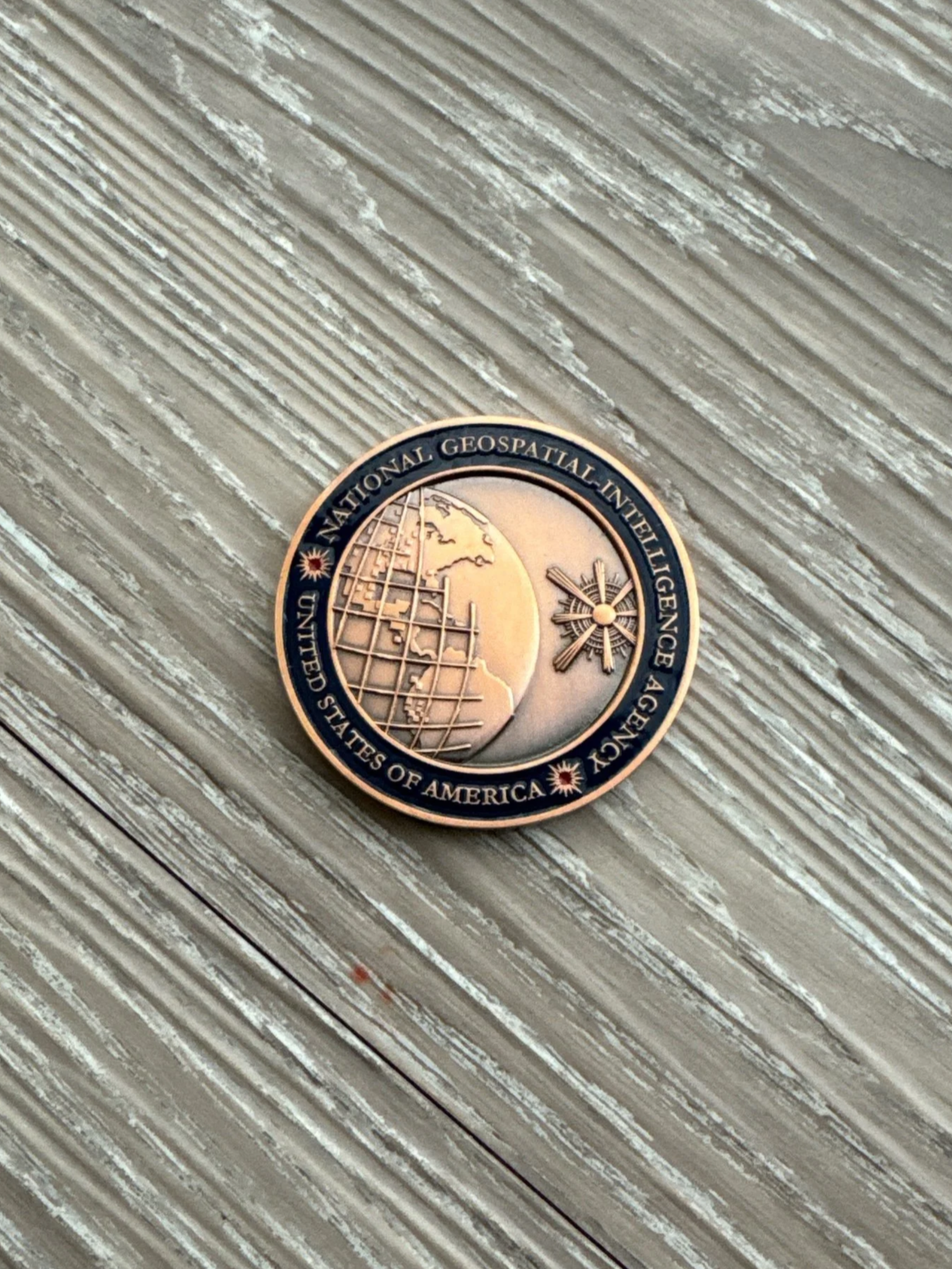 A commemorative coin with the inscription 'National Geospatial Intelligence Agency, United States of America' and a globe with grid lines, an aircraft sensor, and a compass rose, lying on a textured wooden surface.