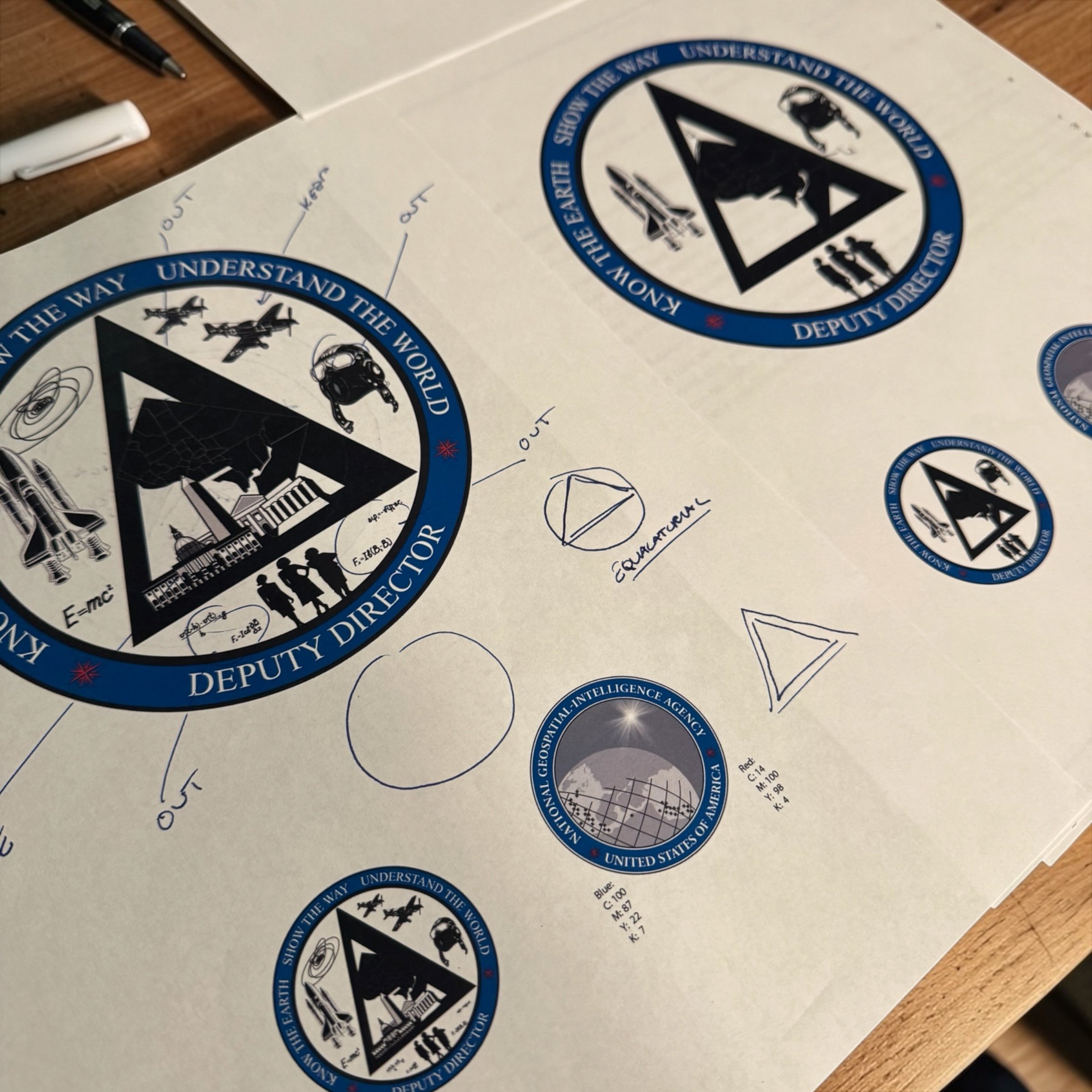 A sheet of paper with various NGA and space-related stickers, including the NGA Deputy Director and National Geospatial-Intelligence Agency logos, along with handwritten notes and diagrams.