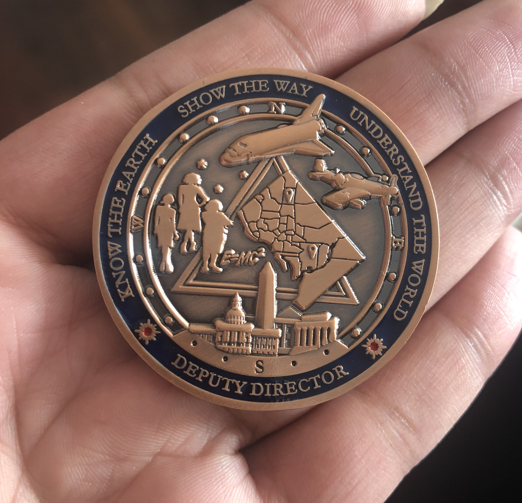 Close-up of a copper-colored medal featuring images of airplanes, people, and government buildings, with the words 'Know the Earth, Show the Way, Understand the World' around the edge, and 'Deputy Director' at the bottom.