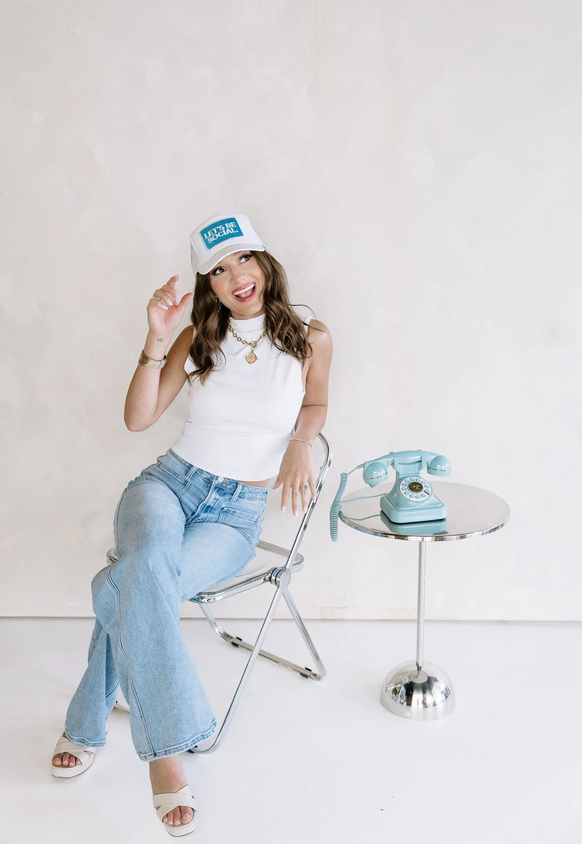 A woman sitting on a chair next to a small round table with a vintage blue rotary phone on it, wearing a white cap and tank top, smiling and holding her hat, in a minimalistic room with a plain light-colored wall.