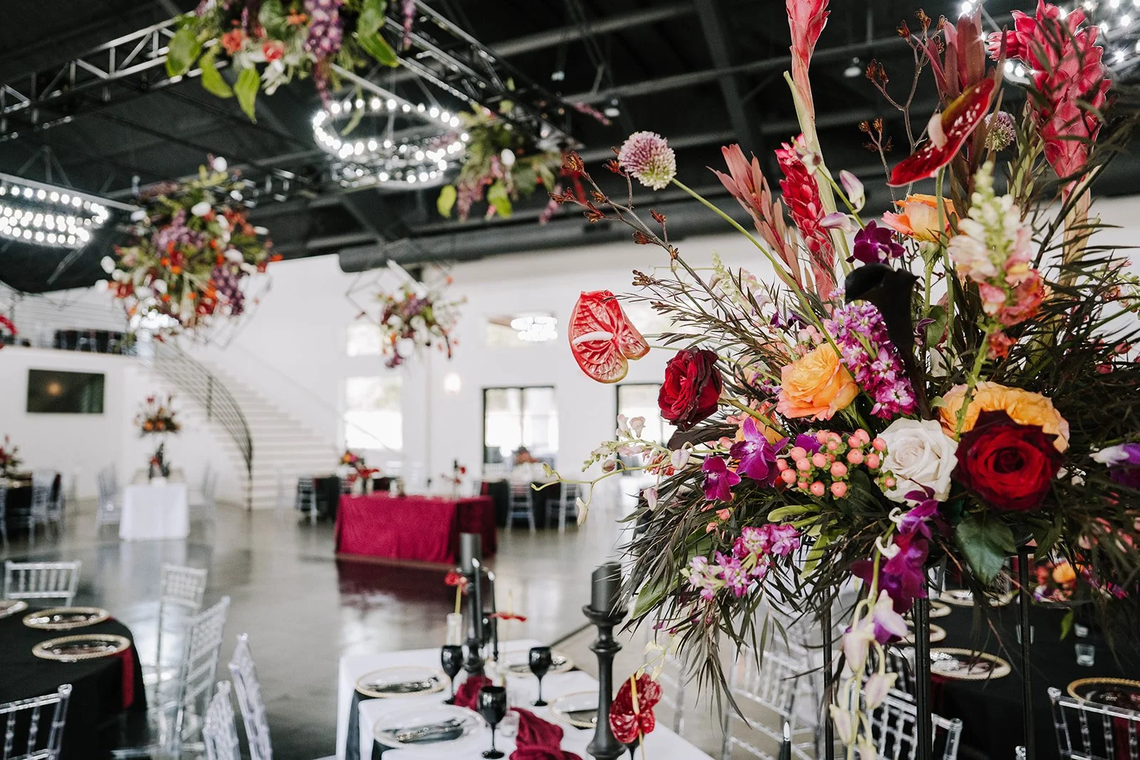 Elegant event venue decorated with floral arrangements, black candles, and tables with black and white table settings, featuring a staircase and modern lighting in the background.