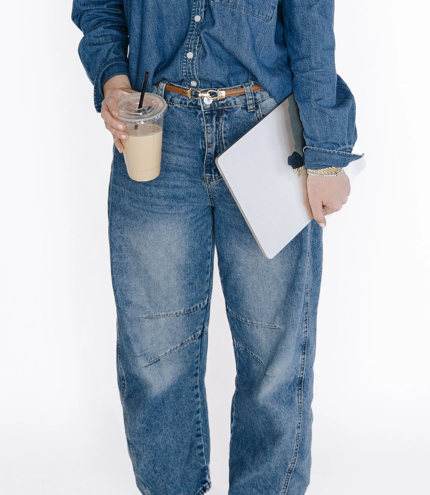 Person wearing a denim shirt and jeans, holding a latte in one hand and a silver laptop in the other, standing against a white background.