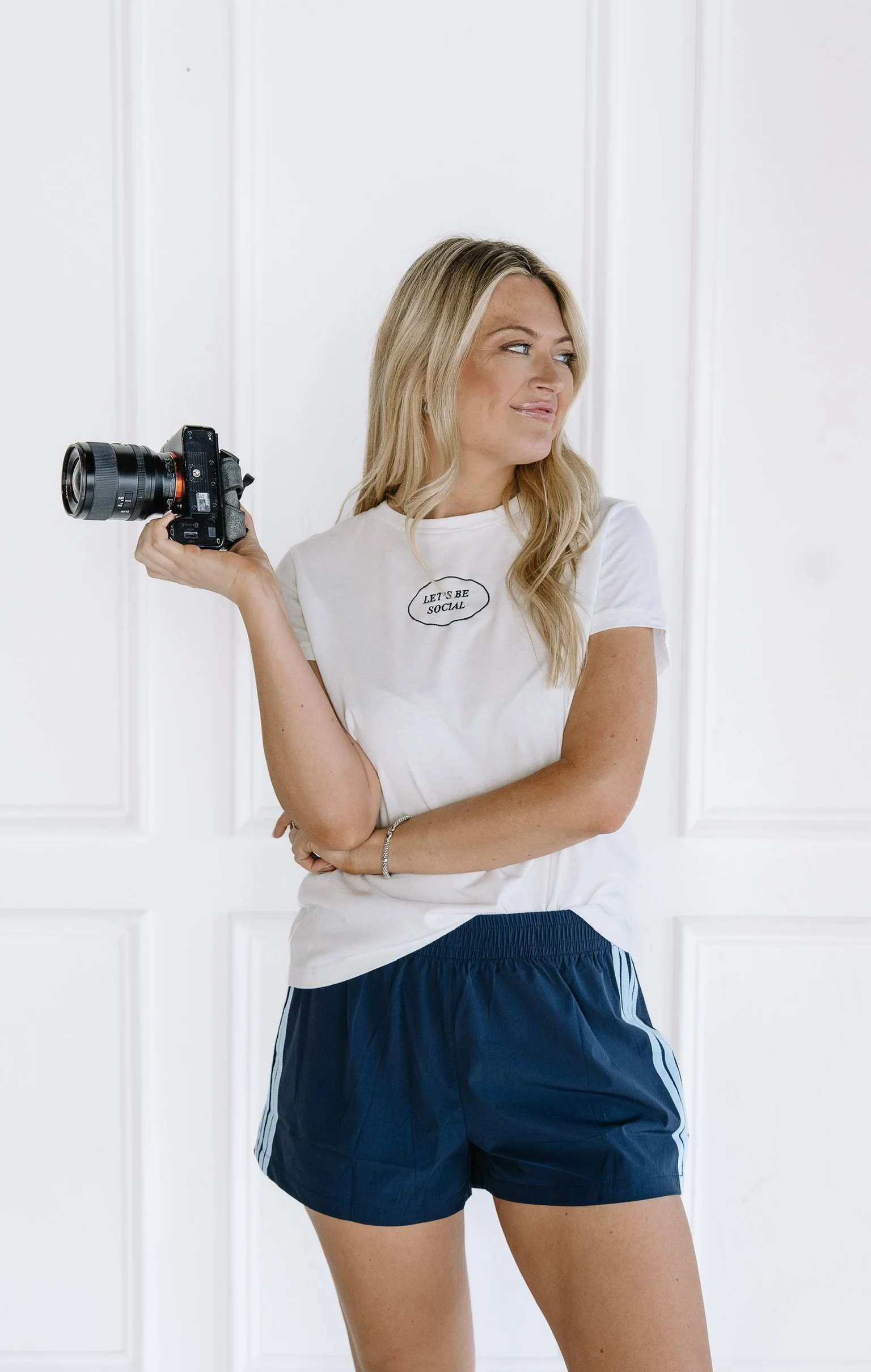 Young woman holding a camera, wearing a white t-shirt with the text 'Let's Be Social' and navy blue athletic shorts, standing in front of a white paneled wall.